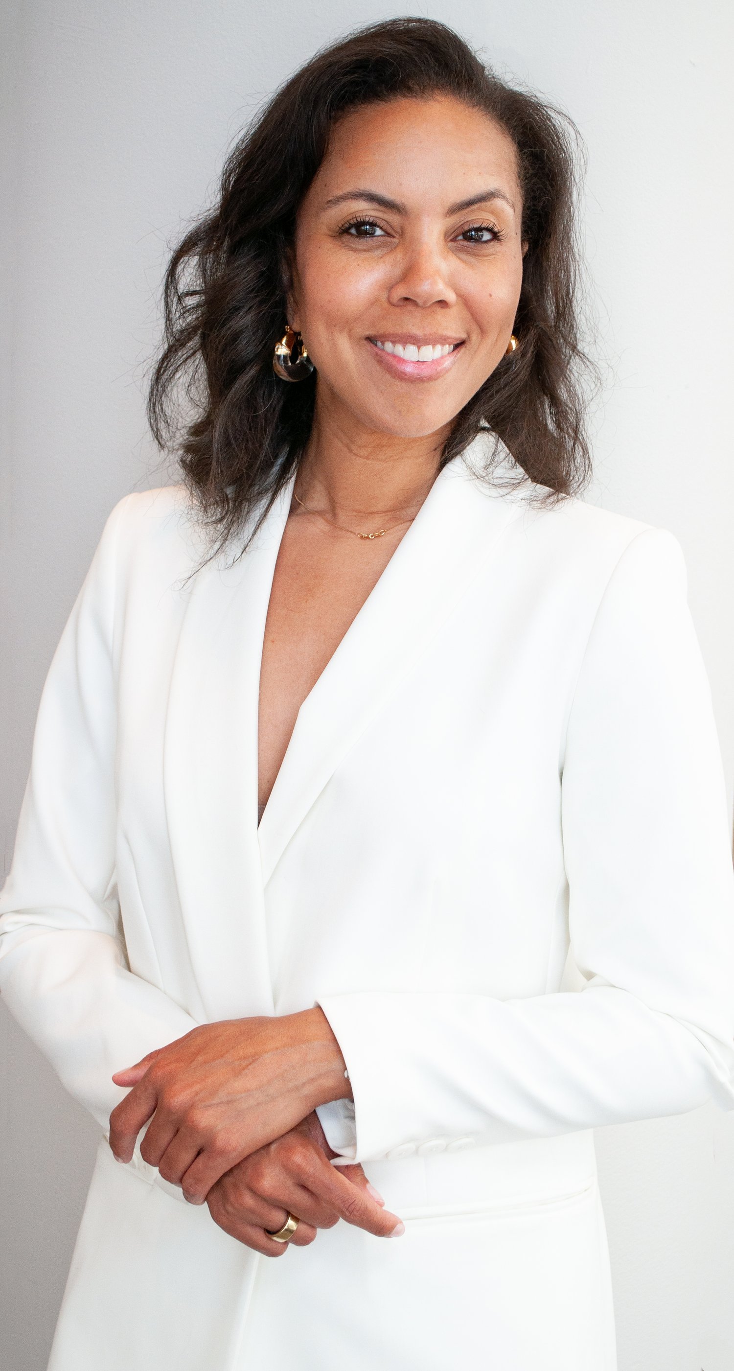 A woman with dark hair and earrings smiling, wearing a brown shirt, sitting on a white cushion or pillow.
