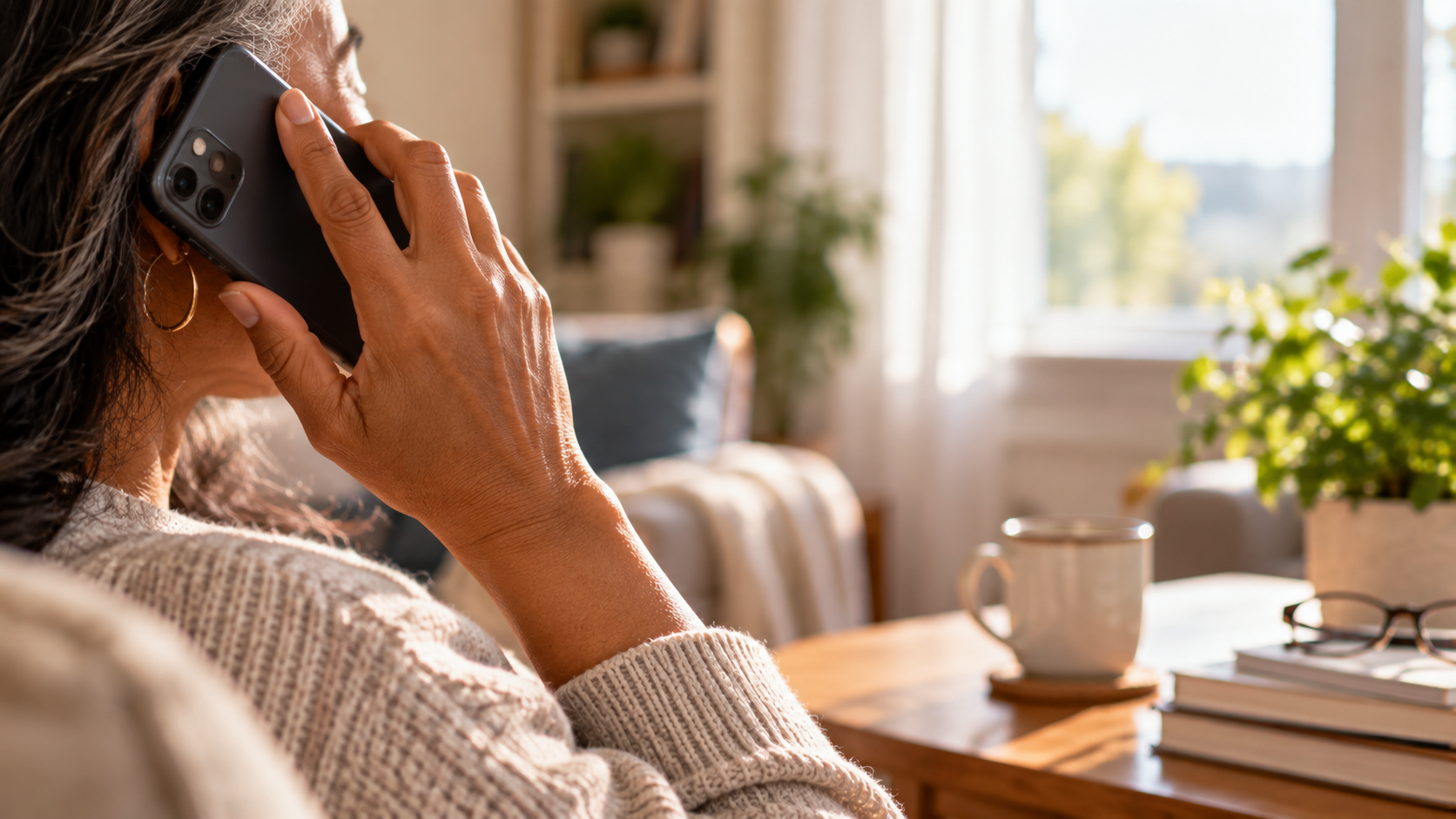 Woman holding a smartphone to her ear during a call in a warm, sunlit home.