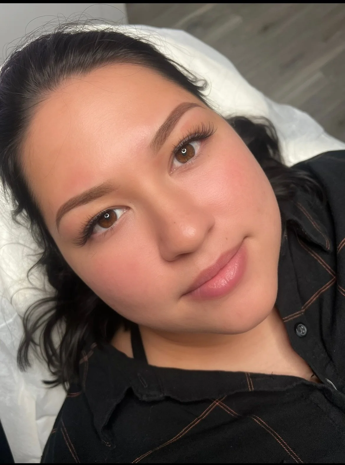 Close-up of a woman lying on a white pillow with makeup, featuring well-groomed eyebrows, long eyelashes, and a slight smile.