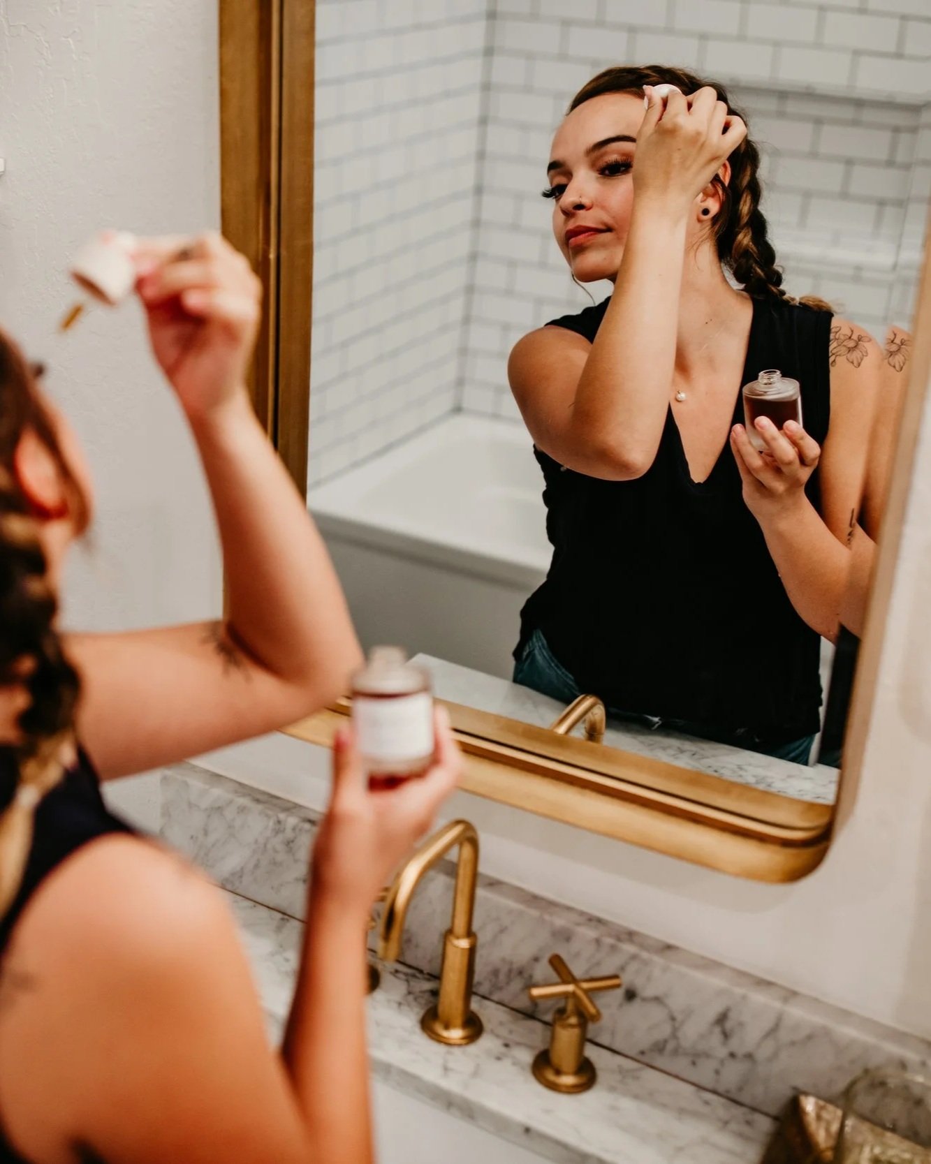 A woman applying skincare cream in front of a mirror in a bathroom, holding a jar of cream in one hand and a spatula in the other. The bathroom has a marble countertop, gold fixtures, and white brick walls.