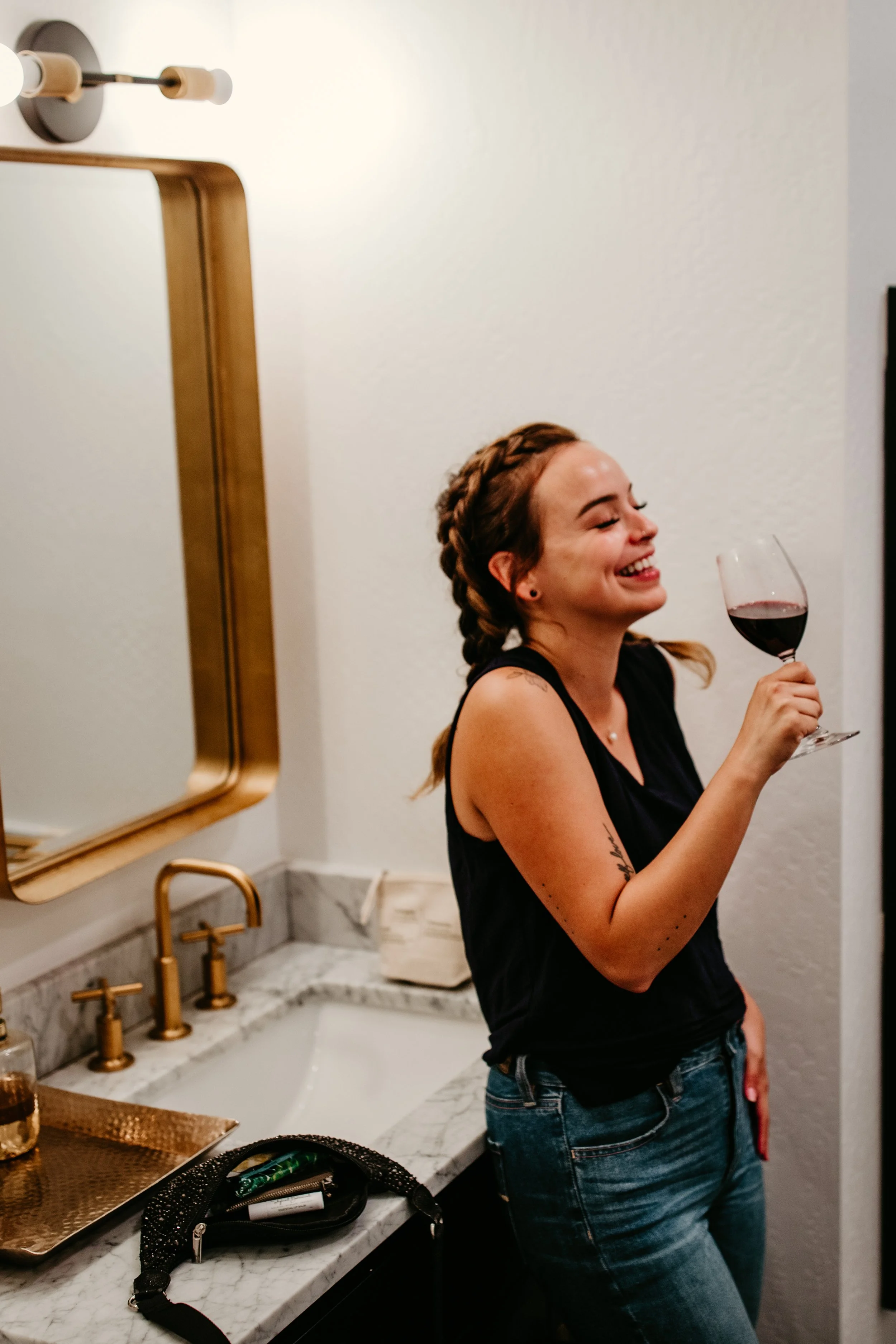 A woman smiling and holding a glass of red wine in a bathroom with gold fixtures, a marble countertop, and a mirror.