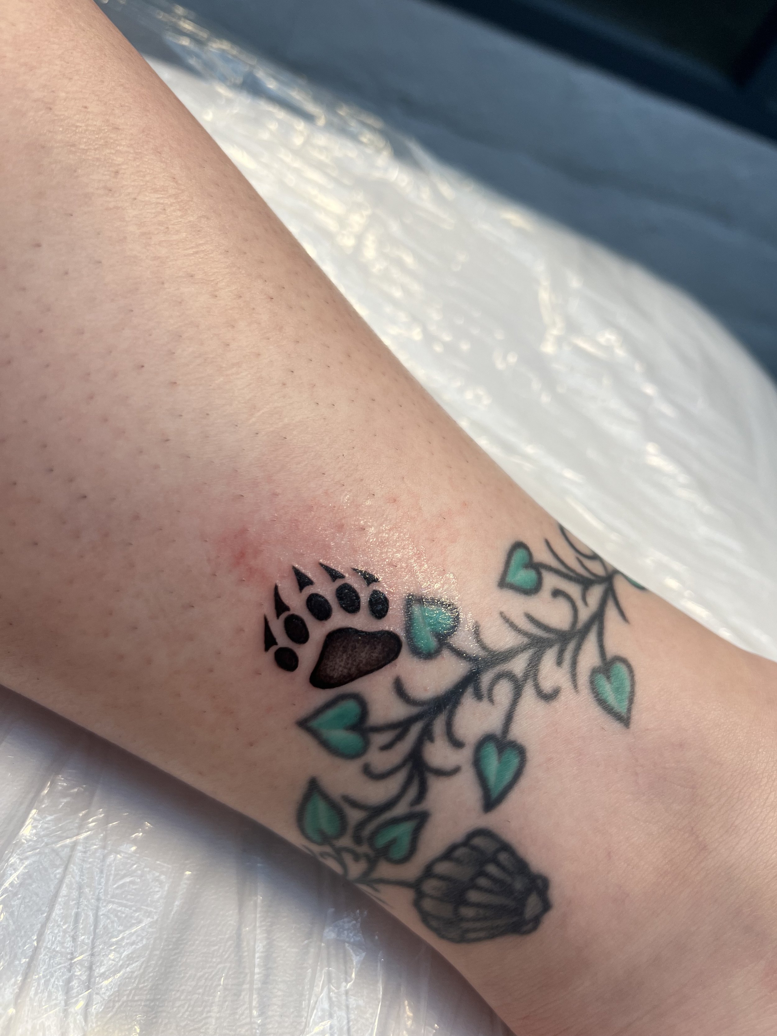 A close-up of a tattoo on a person's forearm featuring a black and gray skull with blue hearts and green foliage.