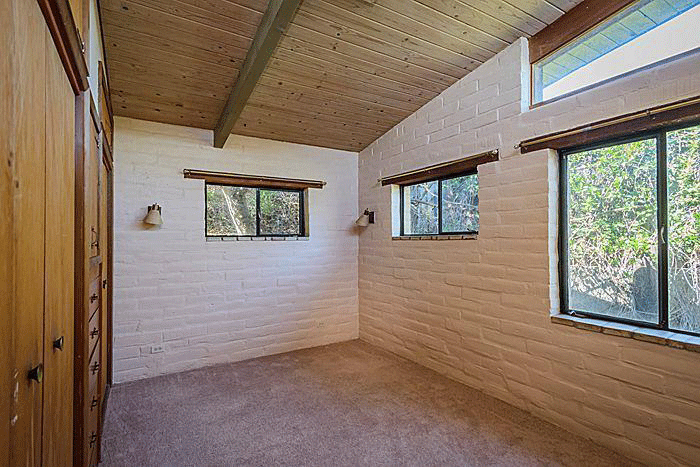 A bedroom of the existing property with high windows, carpet wood ceiling beams and white brick walls