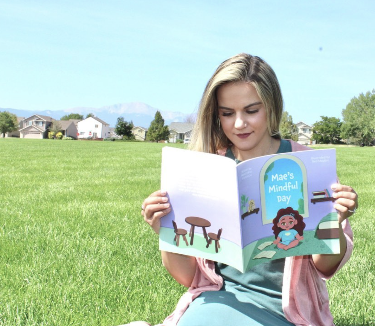 Young woman with blond hair reading booklet titled 'Mae's Mindful Day' outdoors in a grassy park with houses and mountains in the background.