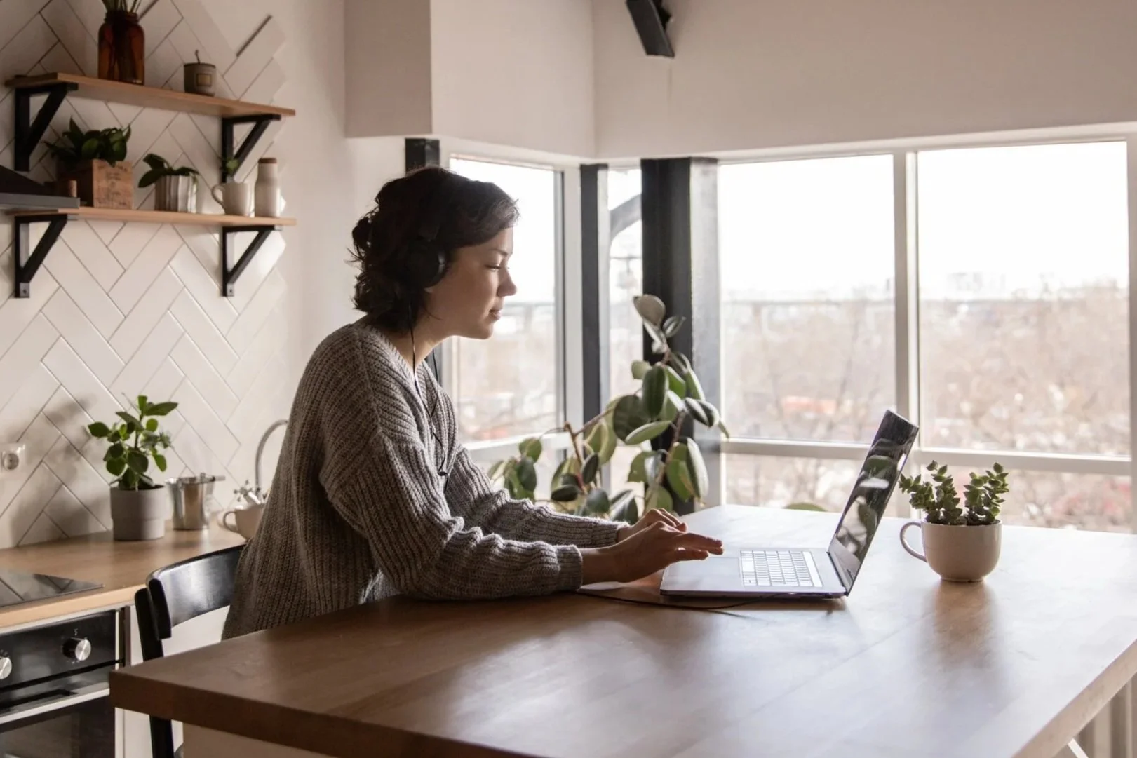 Woman sitting at a kitchen counter using a laptop, wearing headphones, with potted plants and shelves in the background.