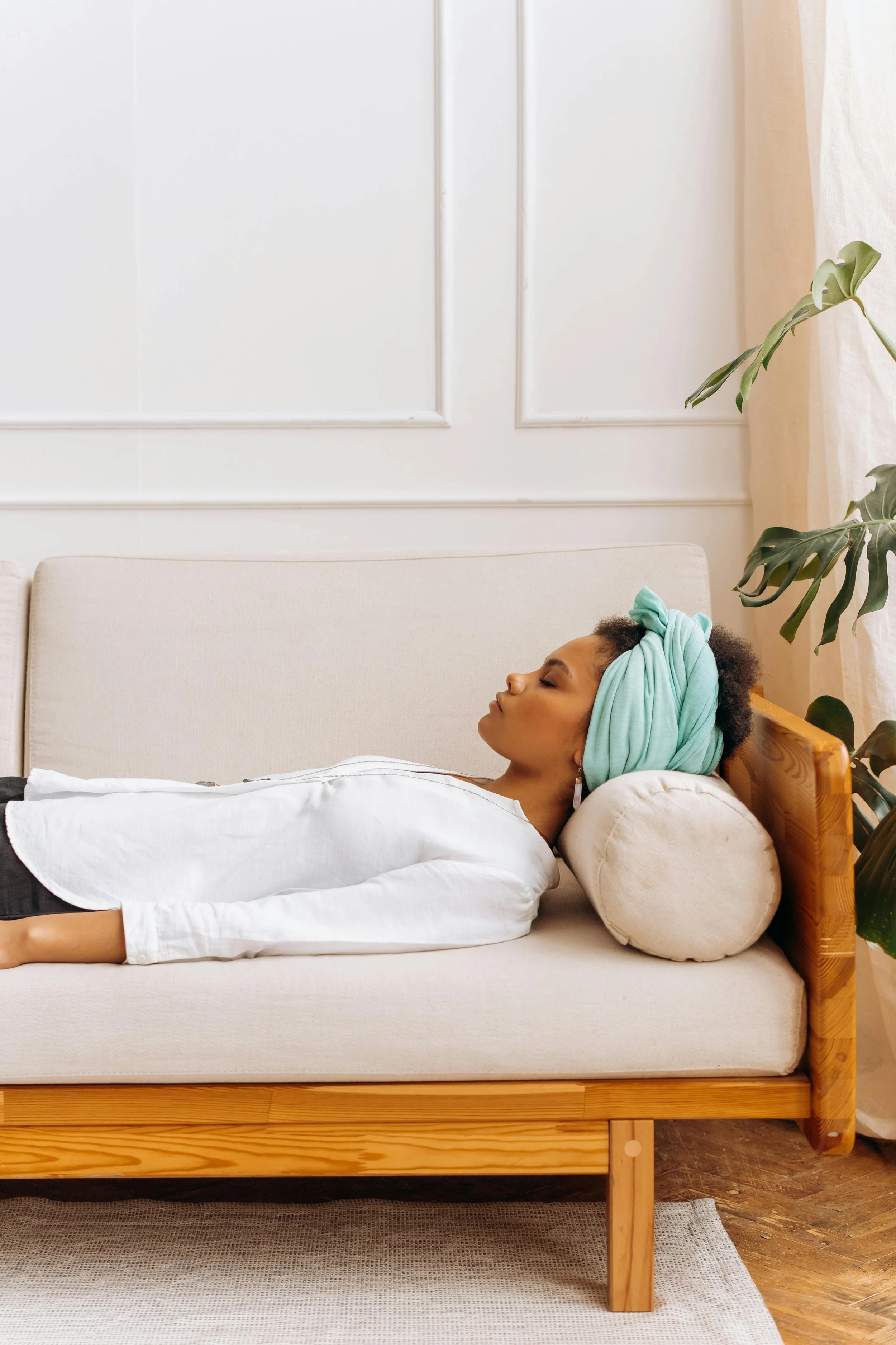 A woman relaxing on a beige sofa with a pillow supporting her head, wearing a turquoise headwrap and a white top, in a bright, minimalist room with white walls and a large plant nearby.