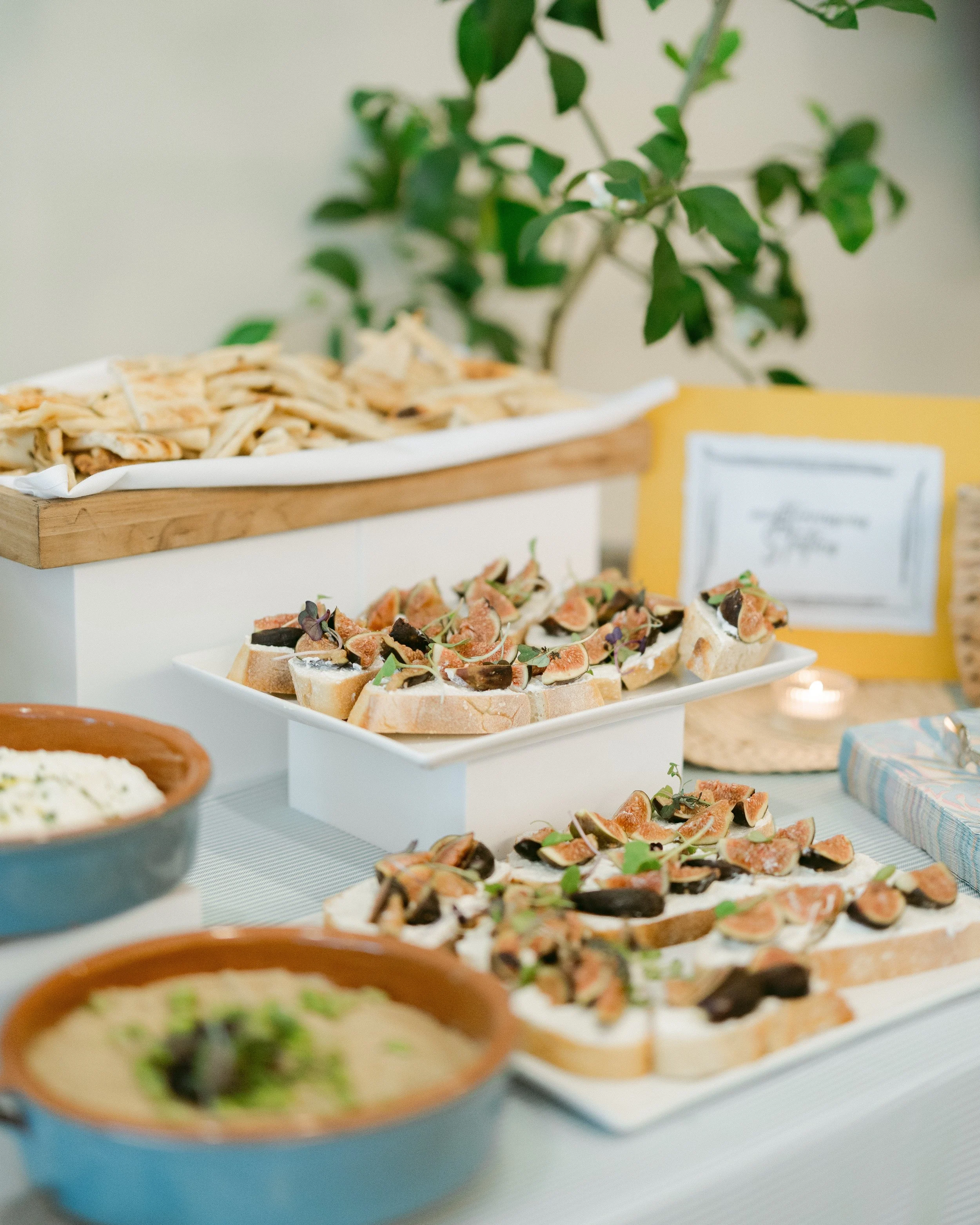 A table with various appetizers and snacks, including bruschetta topped with tomatoes and herbs, and bowls of guacamole and sliced bread, with a blurred background featuring a plant and a yellow sign.