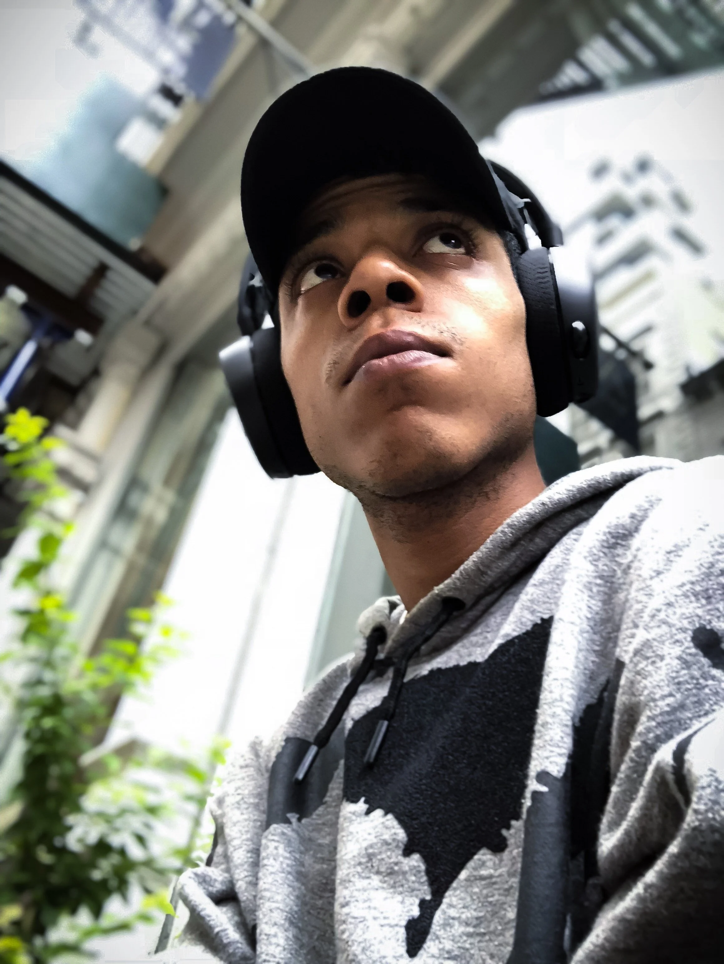 Person wearing headphones and a gray hoodie, looking upwards, in an urban outdoor setting.