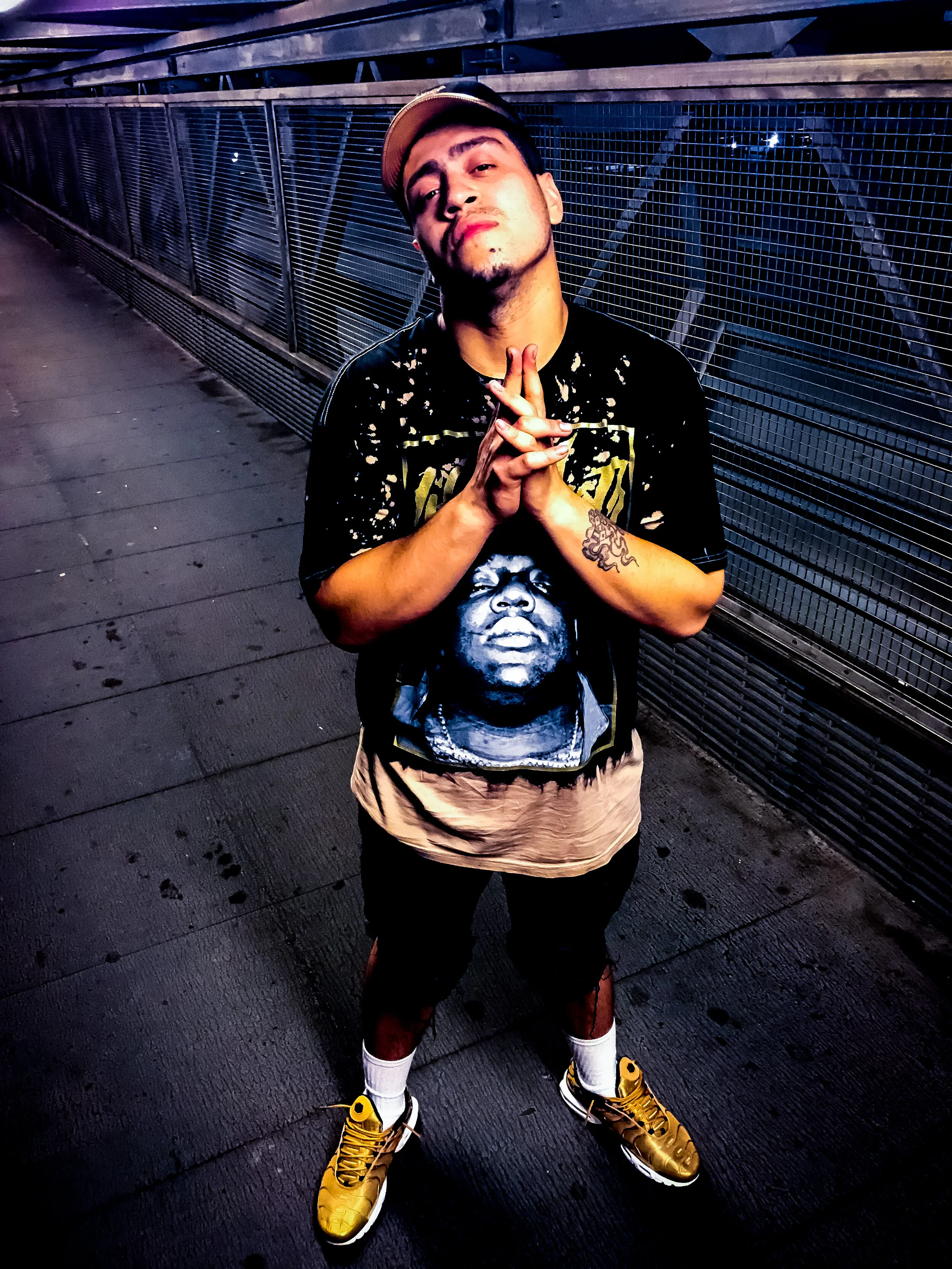 A person in a cap and printed shirt standing on an urban metal walkway with hands clasped, wearing yellow sneakers.