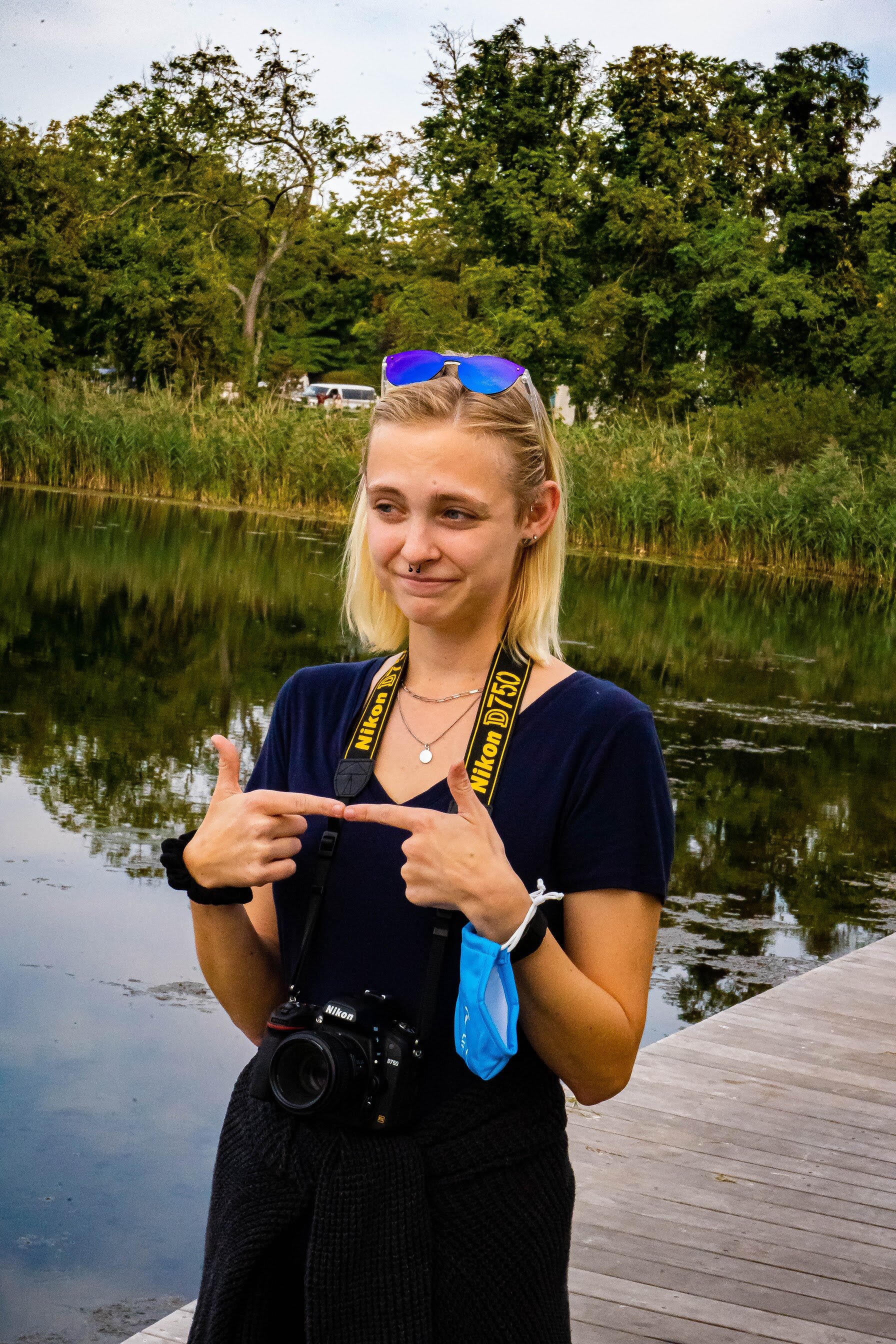 Person with a camera around their neck standing on a wooden path by a lake, pointing at themselves with both hands, wearing sunglasses on their head.