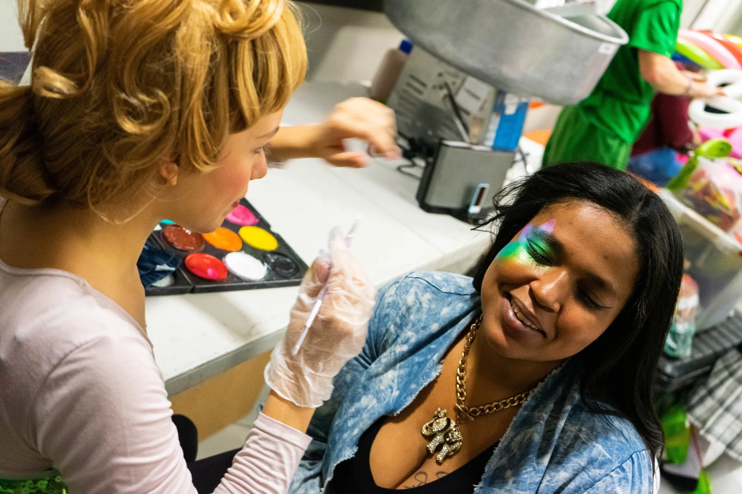 A woman getting face painted with vibrant colors by another person wearing gloves and holding a brush, near a table with paint palette and equipment.