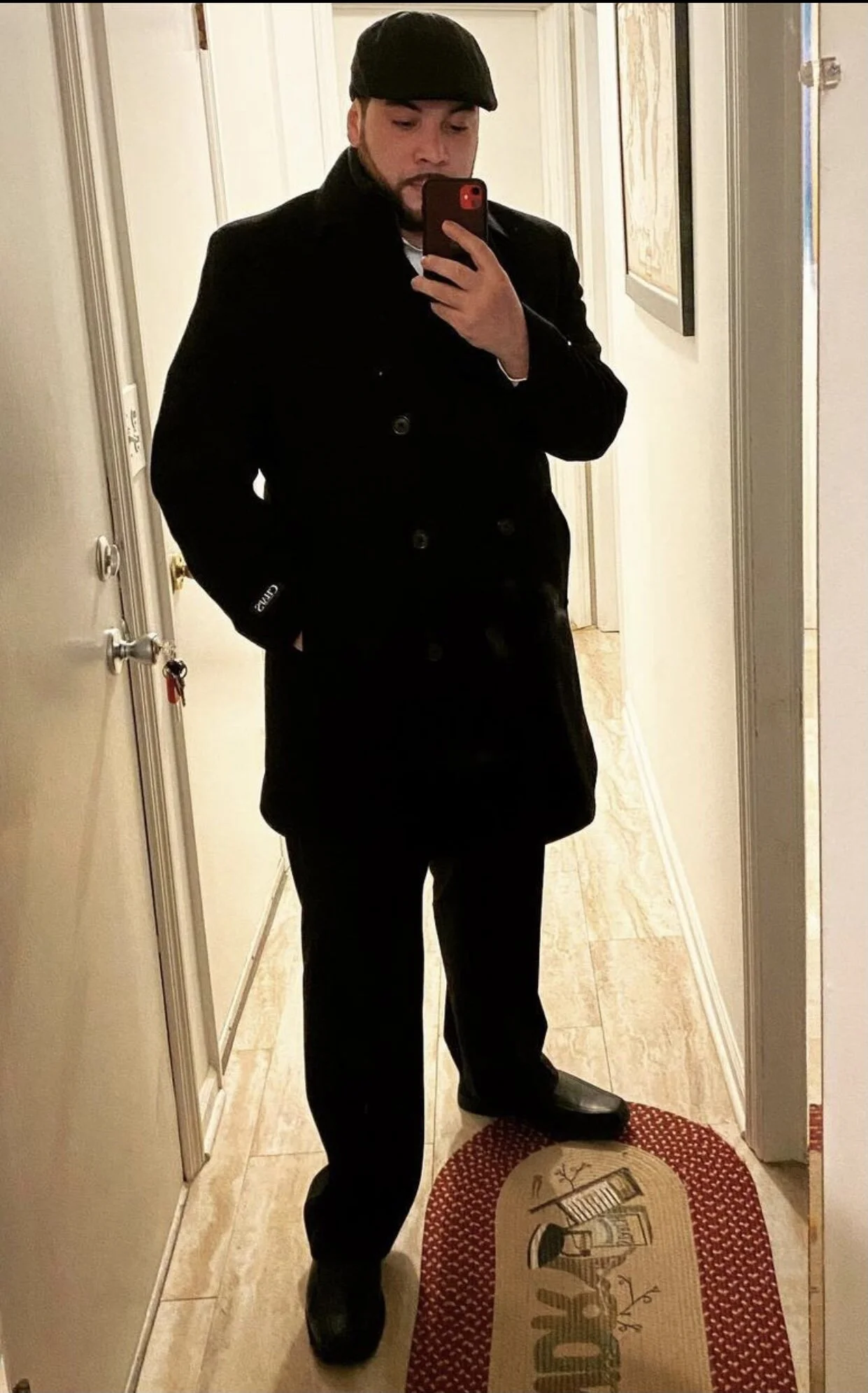 Person taking mirror selfie in hallway, wearing black coat and cap, holding smartphone, with decorative floor mat.
