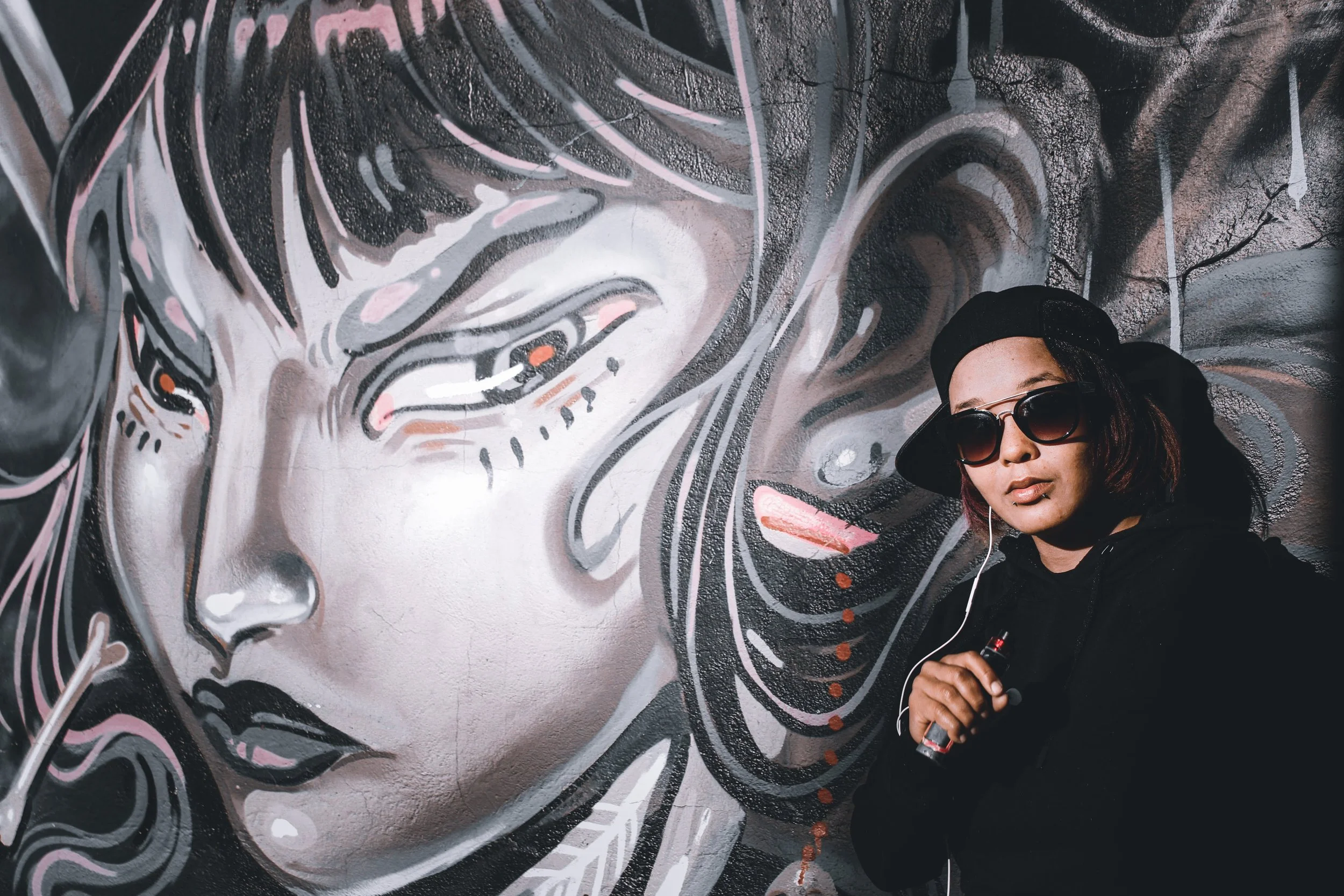 Person wearing sunglasses standing in front of a large, colorful graffiti mural depicting a stylized face on a wall.