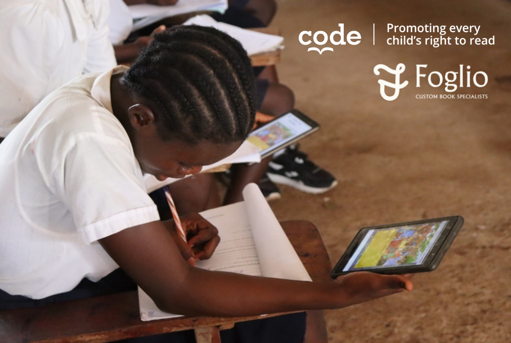 A young student in a white shirt with braids is sitting at a wooden desk in a classroom, holding a tablet device displaying colorful images, with a notebook open in front of her.