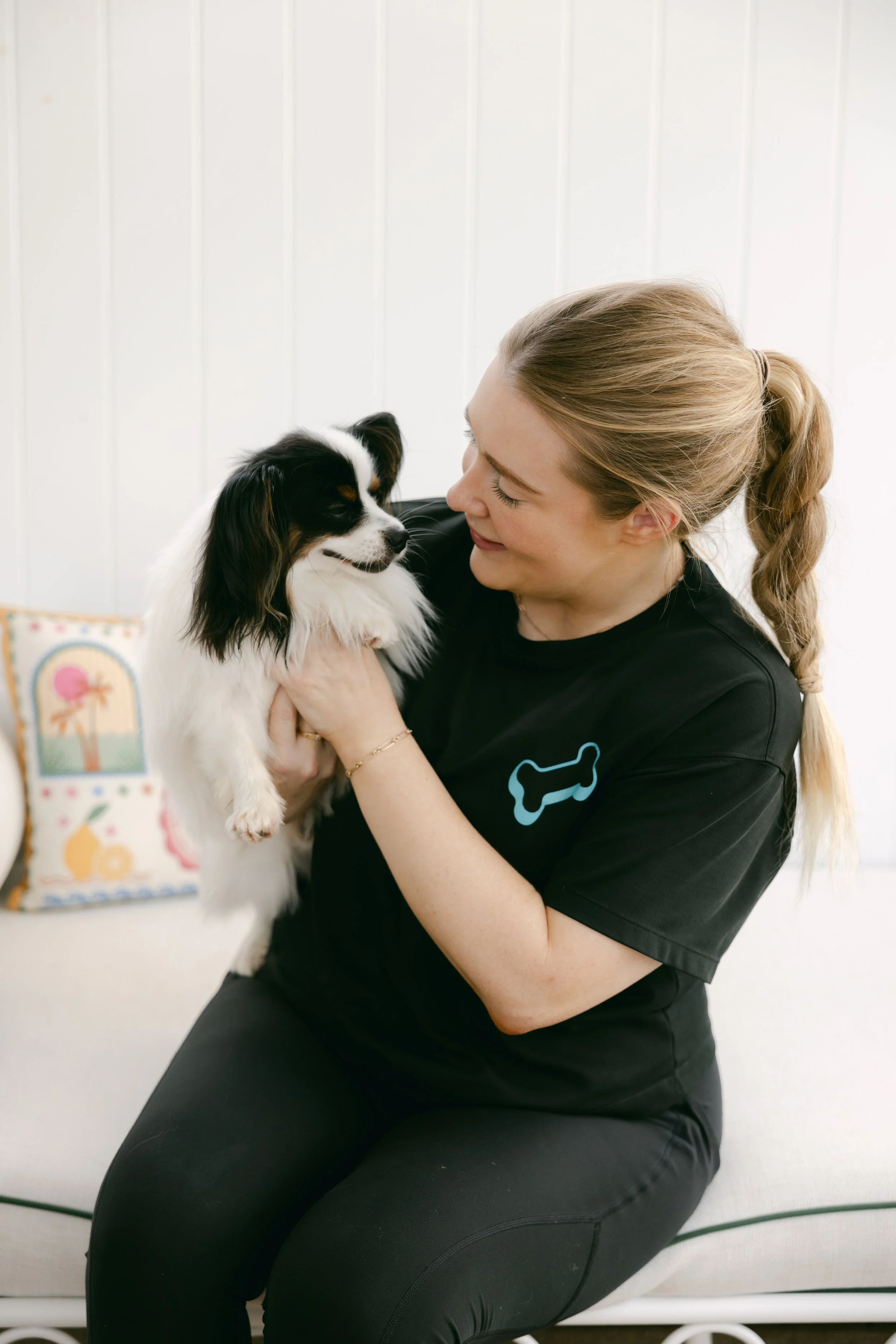 Laura from Doggi smiling while holding a black and white dog, sitting indoors with a white wall in the background.