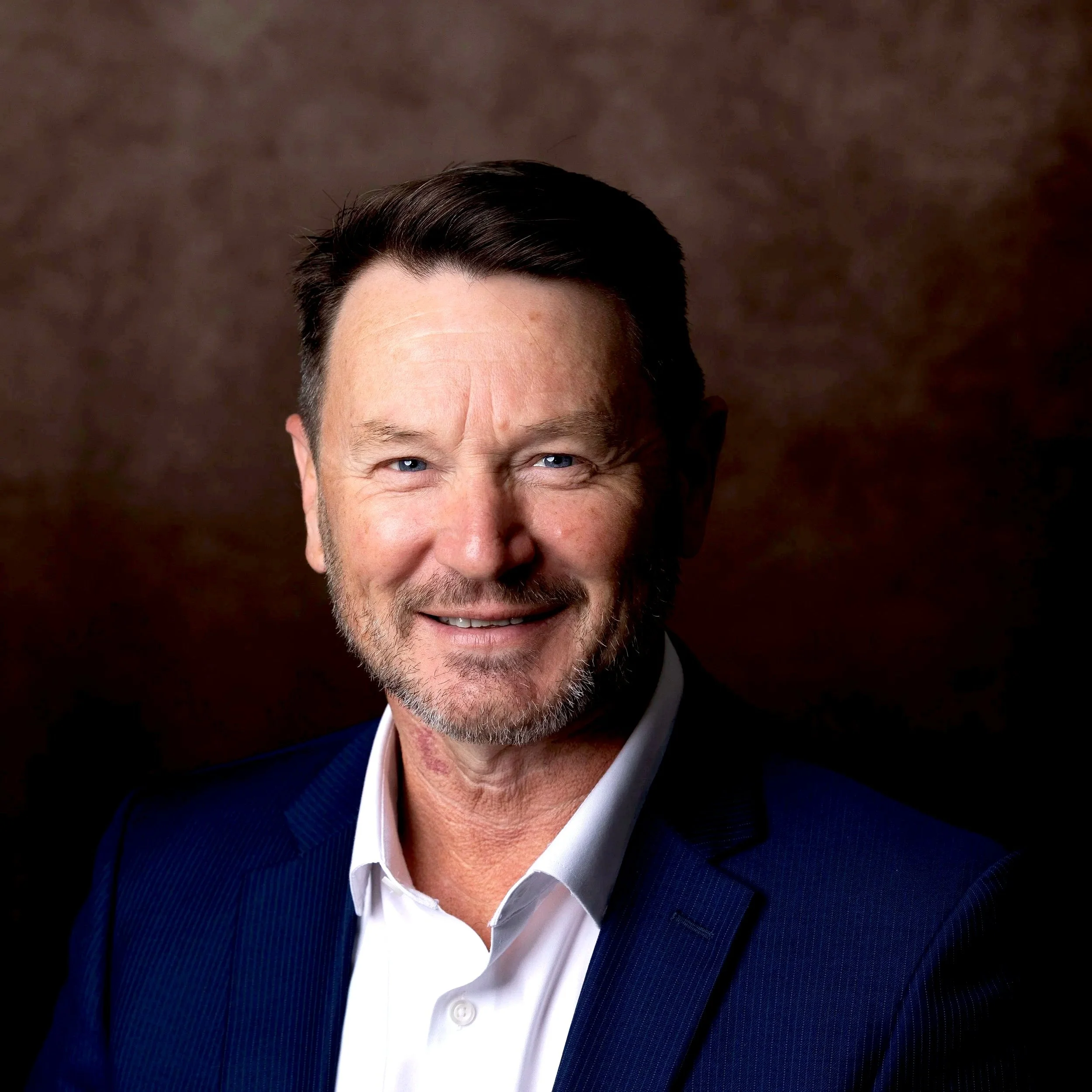 A middle-aged man with dark hair, blue eyes, and a beard, smiling, dressed in a white shirt and blue blazer, against a brown textured background.