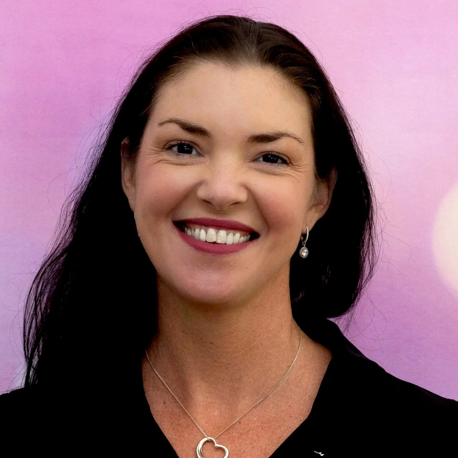 Close-up of a smiling woman with dark hair, wearing earrings and a necklace, against a purple background.