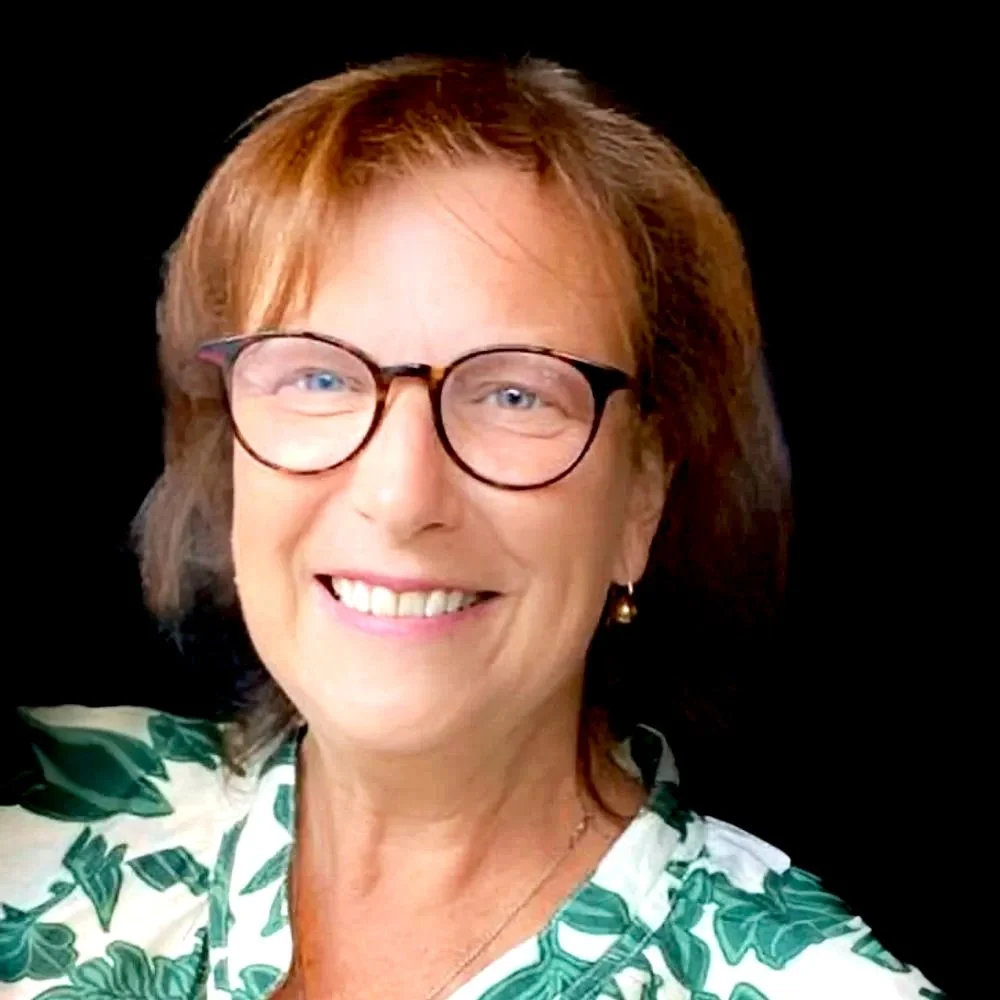 A smiling woman with short, light brown hair, wearing glasses with round frames, a white top with green leaf patterns, gold earrings, and a necklace, sitting in front of a dark background.
