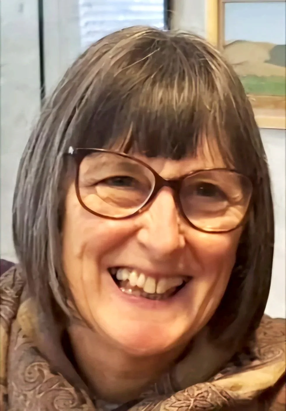 A smiling woman with brown hair, glasses, and a patterned top, indoors by a window and a framed picture.