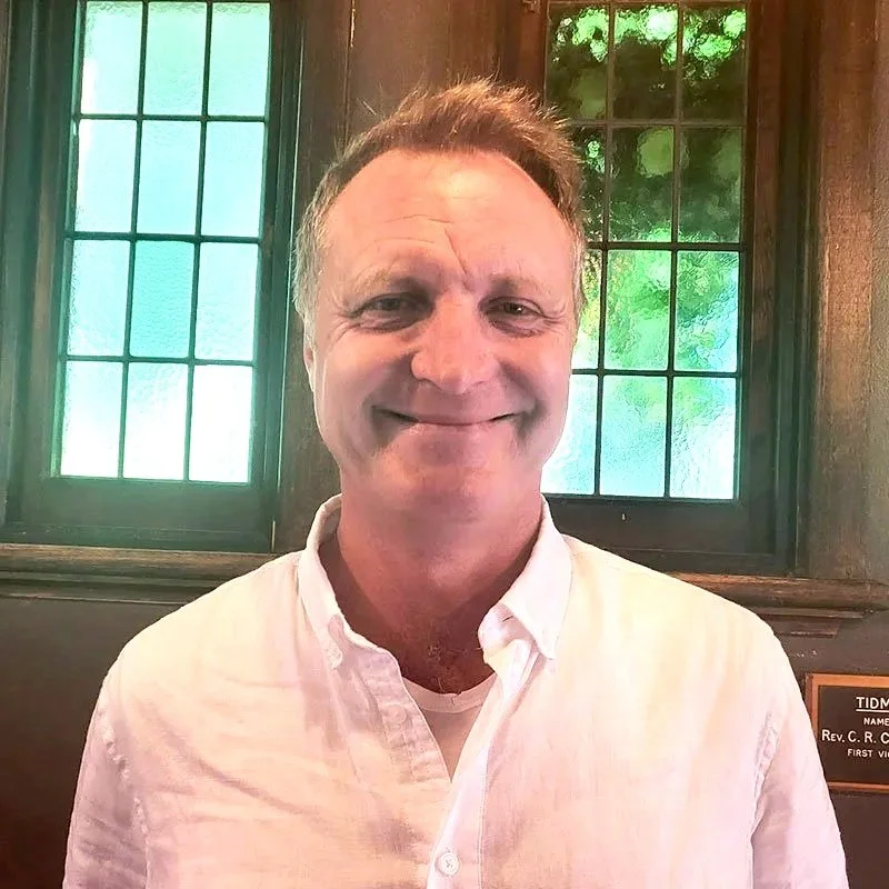 A man smiling in front of decorative stained glass windows.