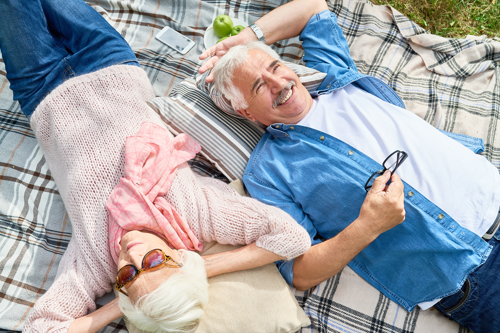 An elderly woman with white hair wearing sunglasses and a pink sweater lying on a picnic blanket, and an elderly man with white hair and a mustache lying on his back on a different blanket, holding glasses and smiling. There are green apples and a smartphone on the blanket near the man.