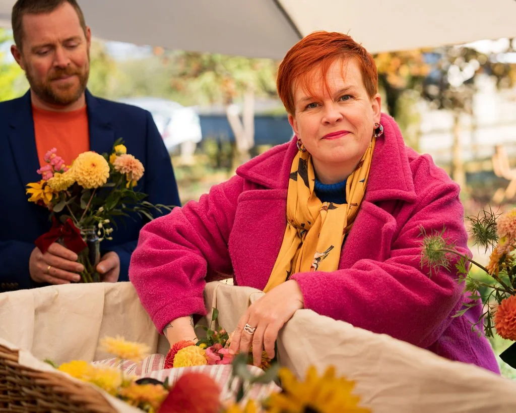 A woman with short bright red hair and wearing a pink coat and yellow scarf smiles into the camera as she places something into an open wicker coffin