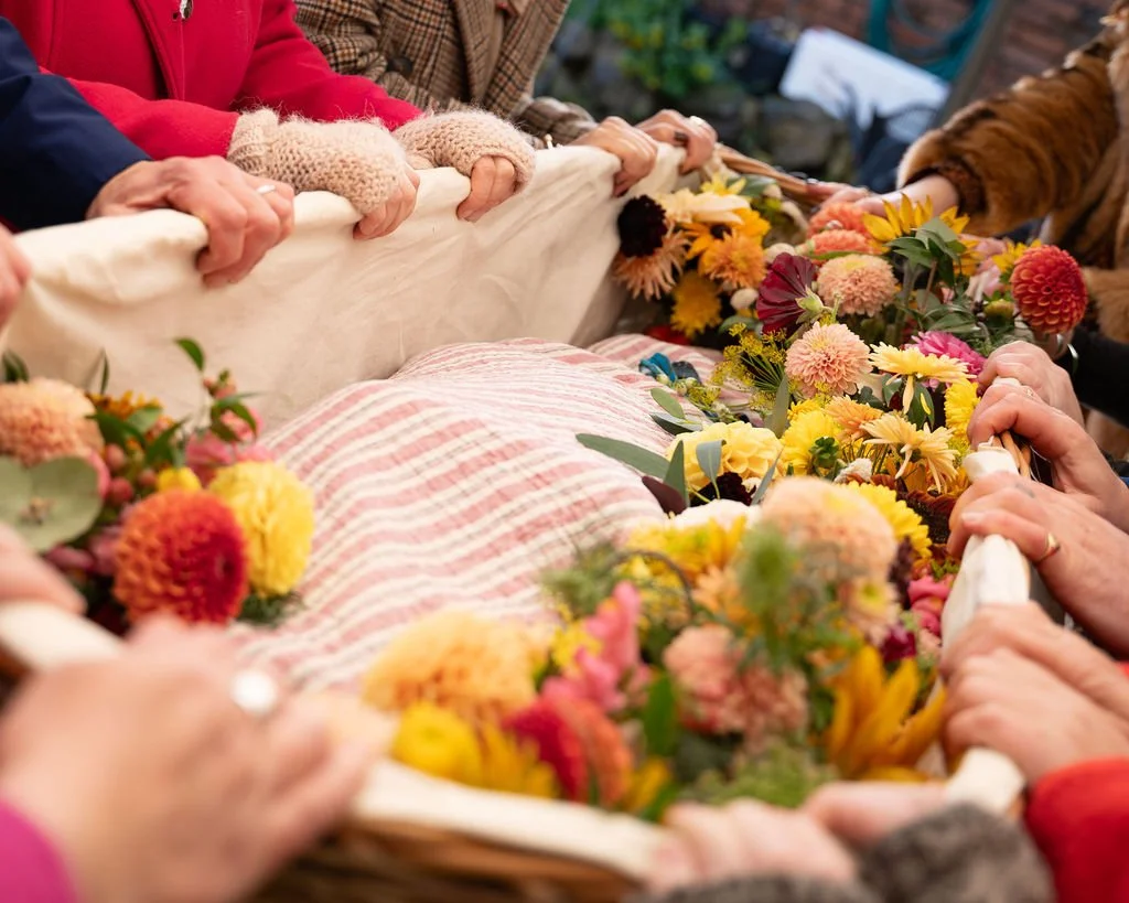 Hands hold onto the edge of a wicker coffin that contains autumnal flowers and a shrouded body