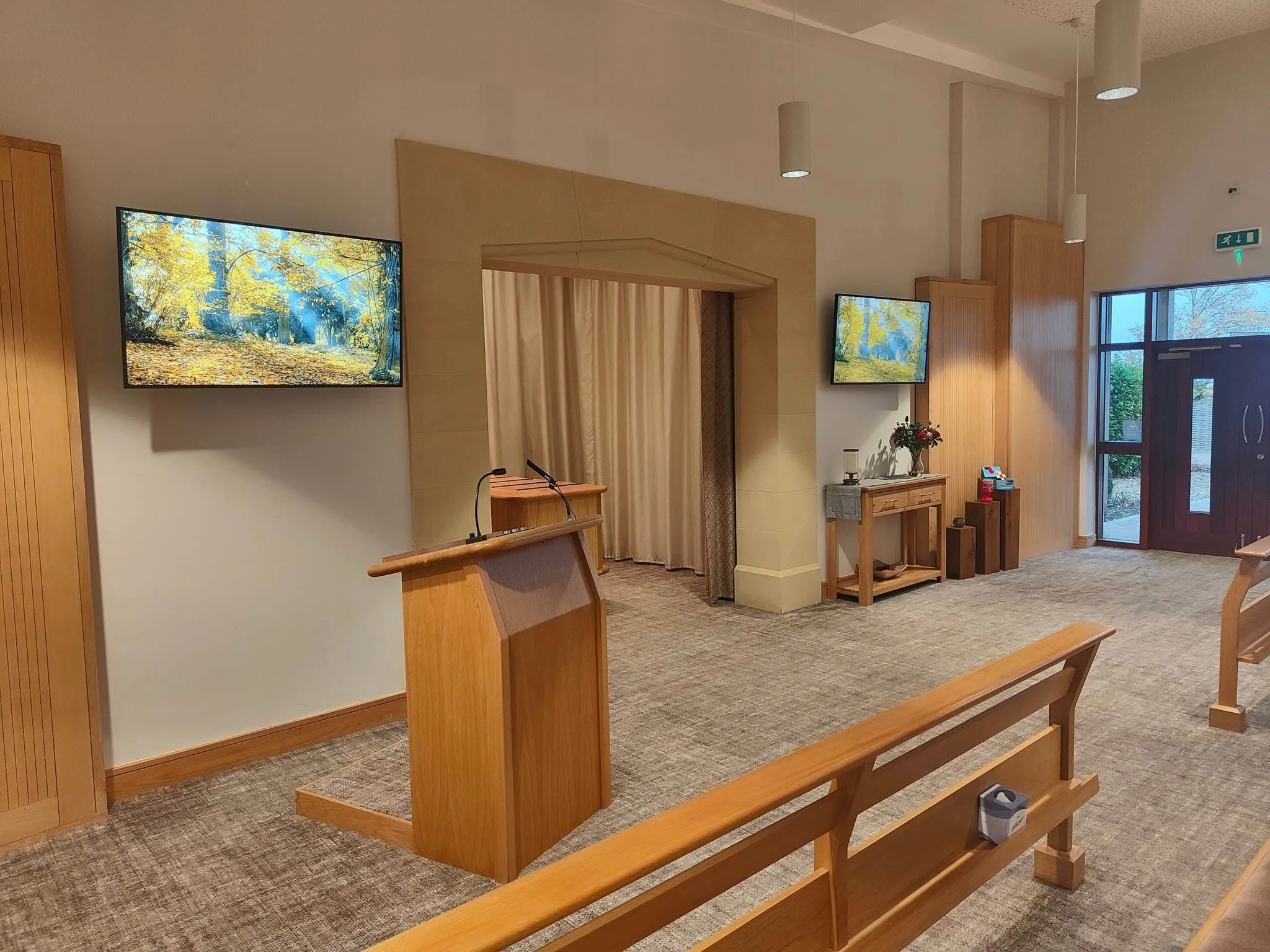 The interior of a crematorium chapel with beige carpet, wooden benches and lectern with attached microphone. Two TV screens are attached to the cream wall behind the lectern, showing an autumnal scene. Inbetween the two screens is an arched opening