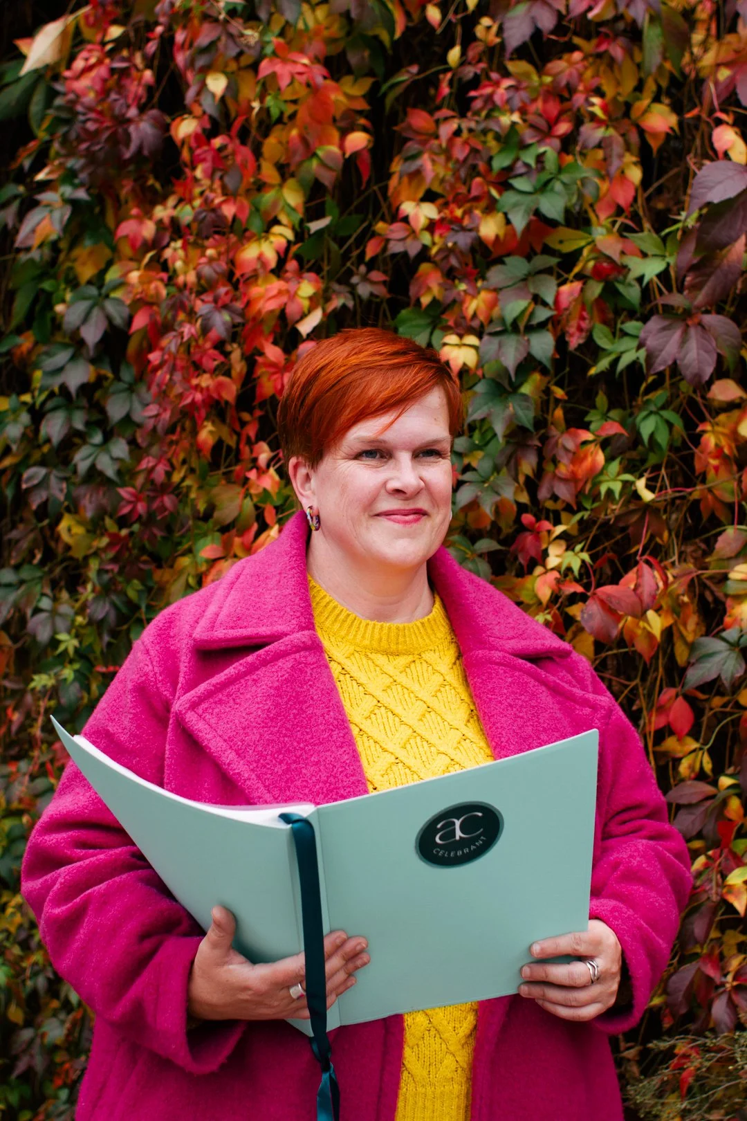 A woman with short bright red hair and wearing a yellow jumper and a bright pink coat stands in front of a wall covered with autumnal creepers and holds a pale turquoise folder open in front of her