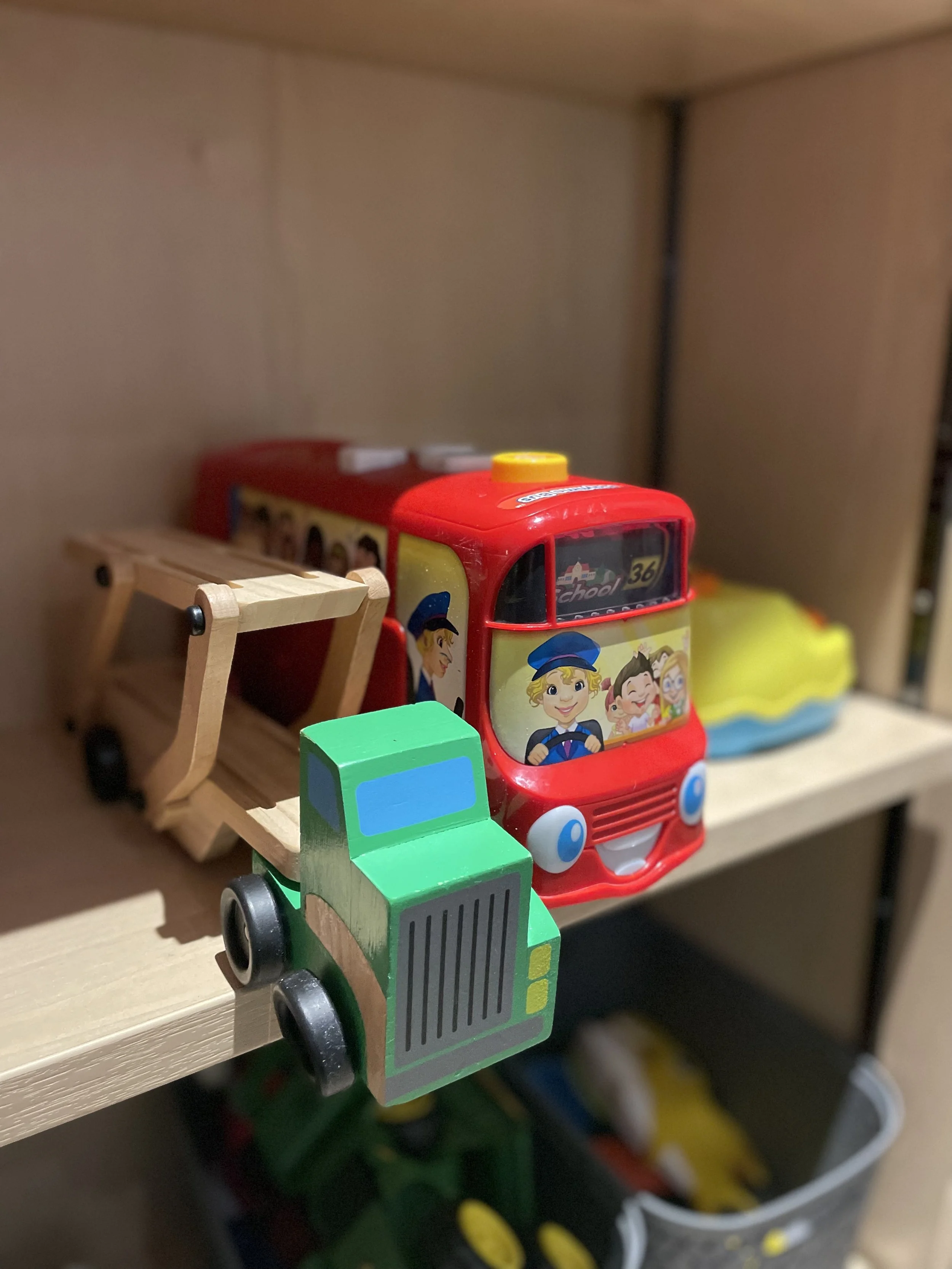 A green wooden toy lorry sits next to a red plastic toy bus on a wooden shelf
