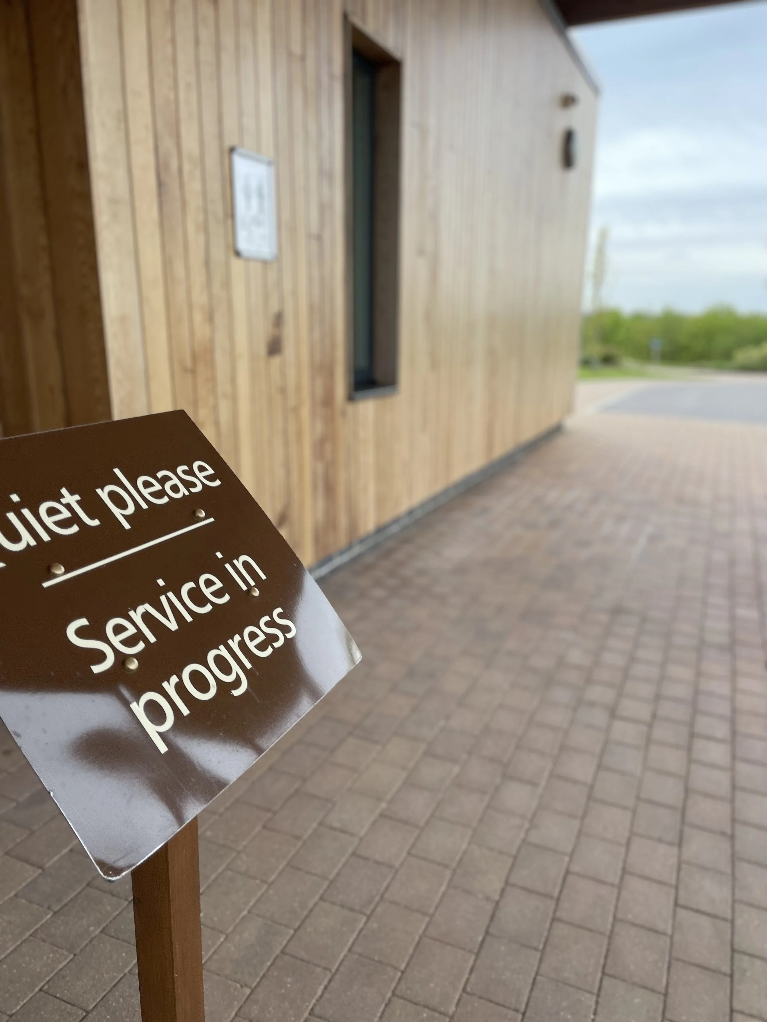 A sign saying Quiet Please Service In Progress stands on a brick floor with a wooden clad building in the background