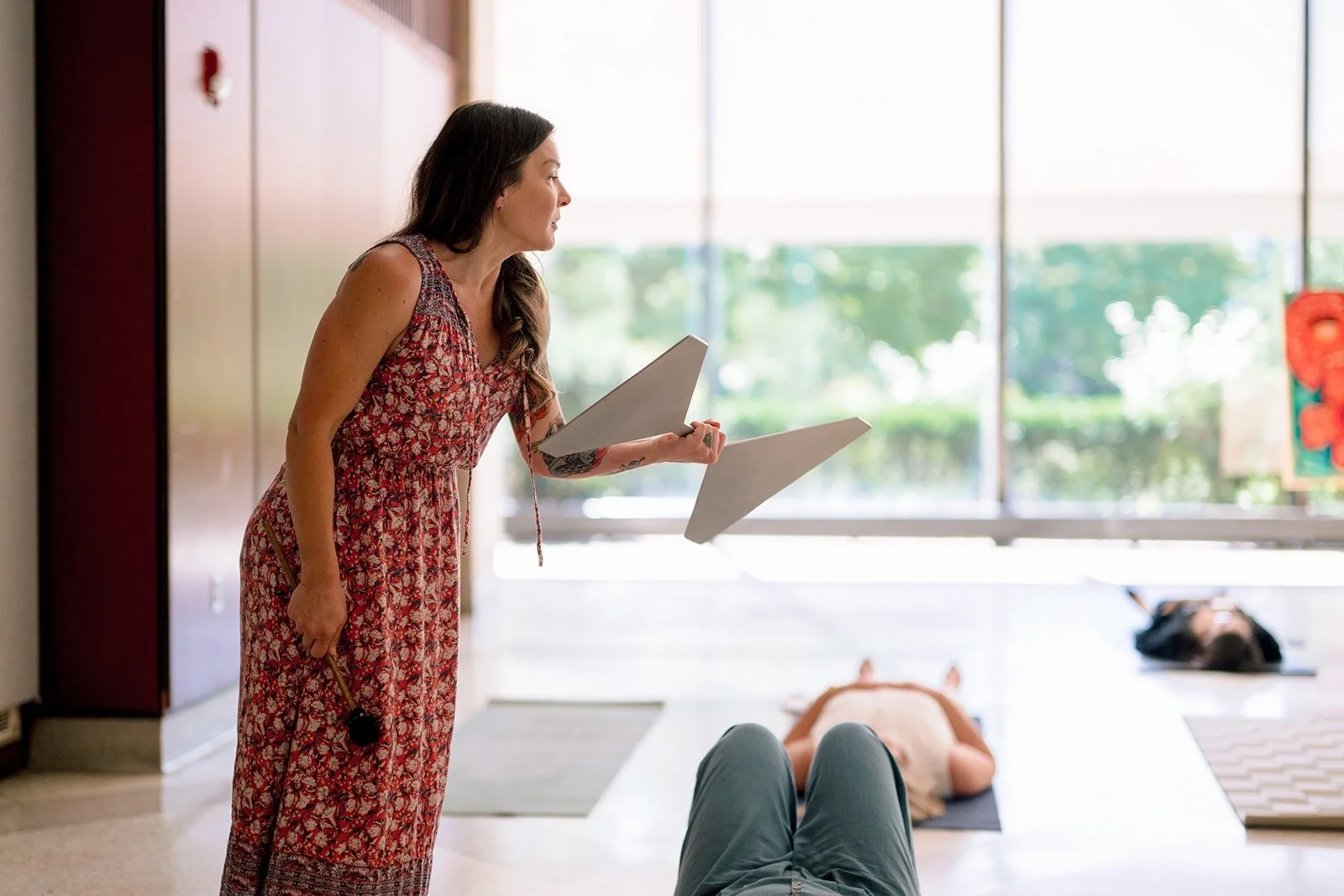 A woman in a floral dress leading a meditation or yoga session in a bright room, with another person lying on a yoga mat and someone else practicing yoga in the background.