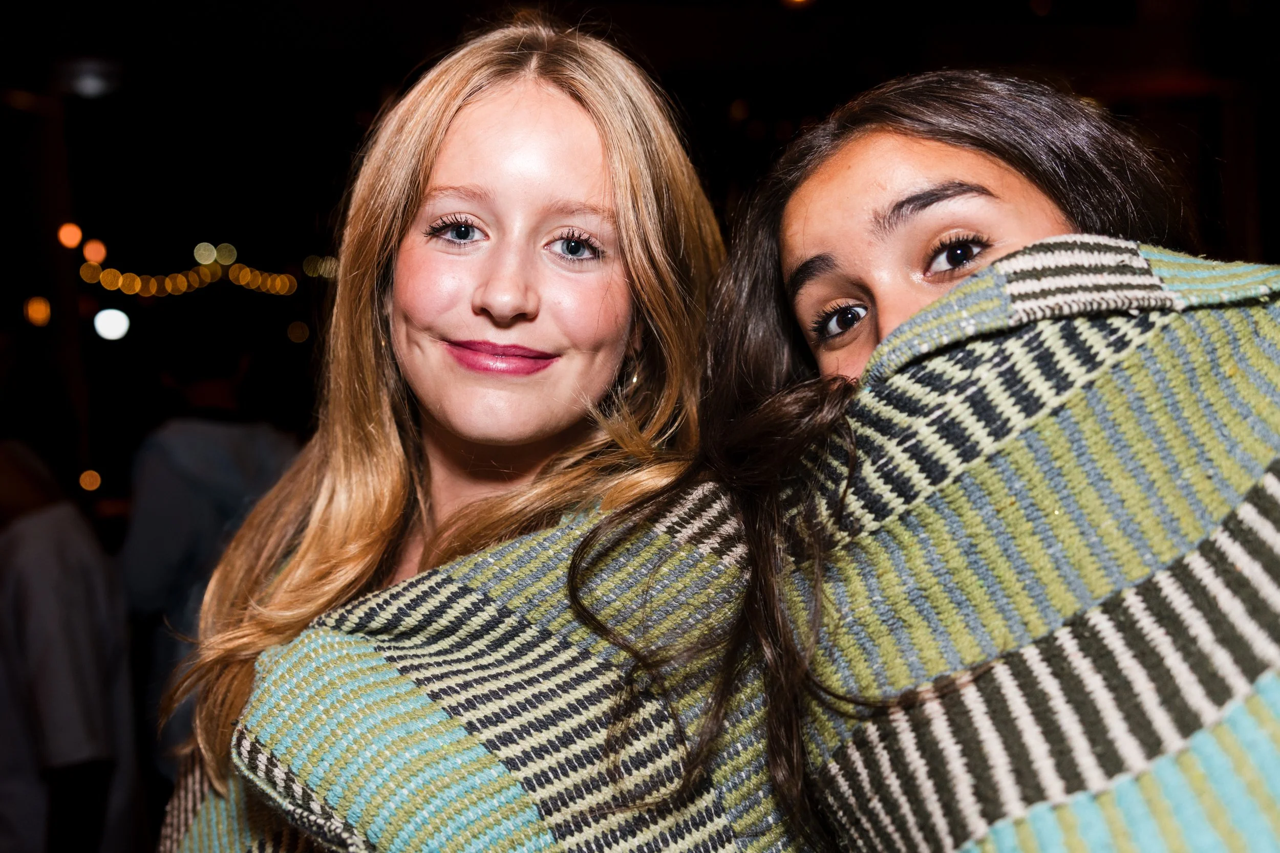 Two young women smiling at the camera, one with blonde hair and the other with dark hair partially covered by a striped jacket, at a bat mitzvah party with string lights in the background.