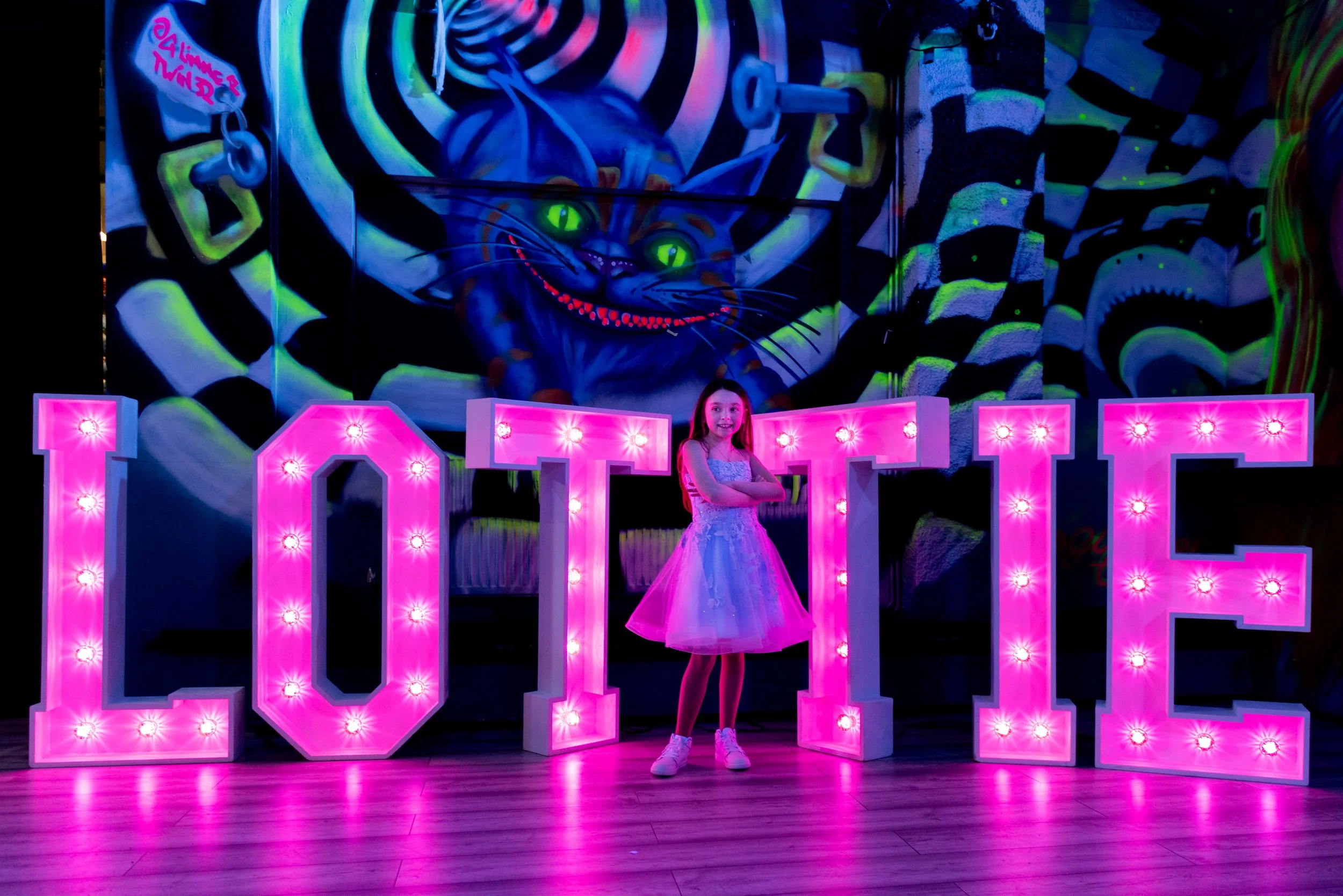 A young girl in a light blue dress stands with her arms crossed in front of large pink illuminated letters spelling 'LOVE' on a wooden floor. 