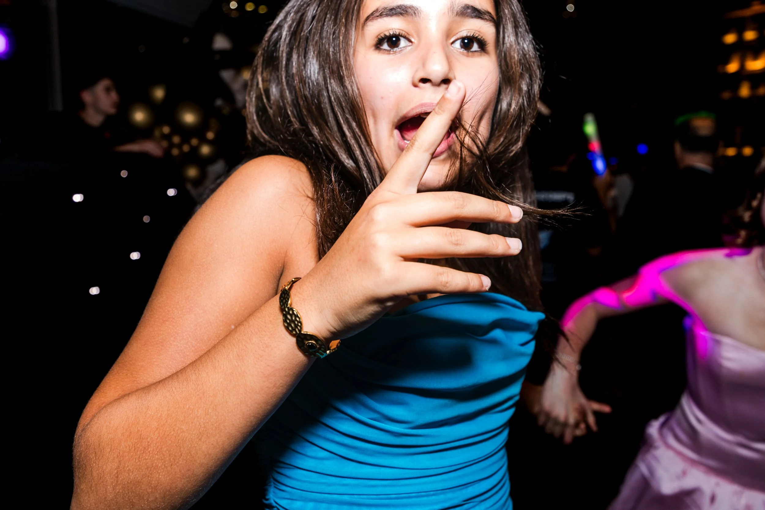 A young woman with brown hair and light skin wearing a strapless blue dress and a black bracelet, holding one finger to her lips in a shushing gesture at a luxurious party.