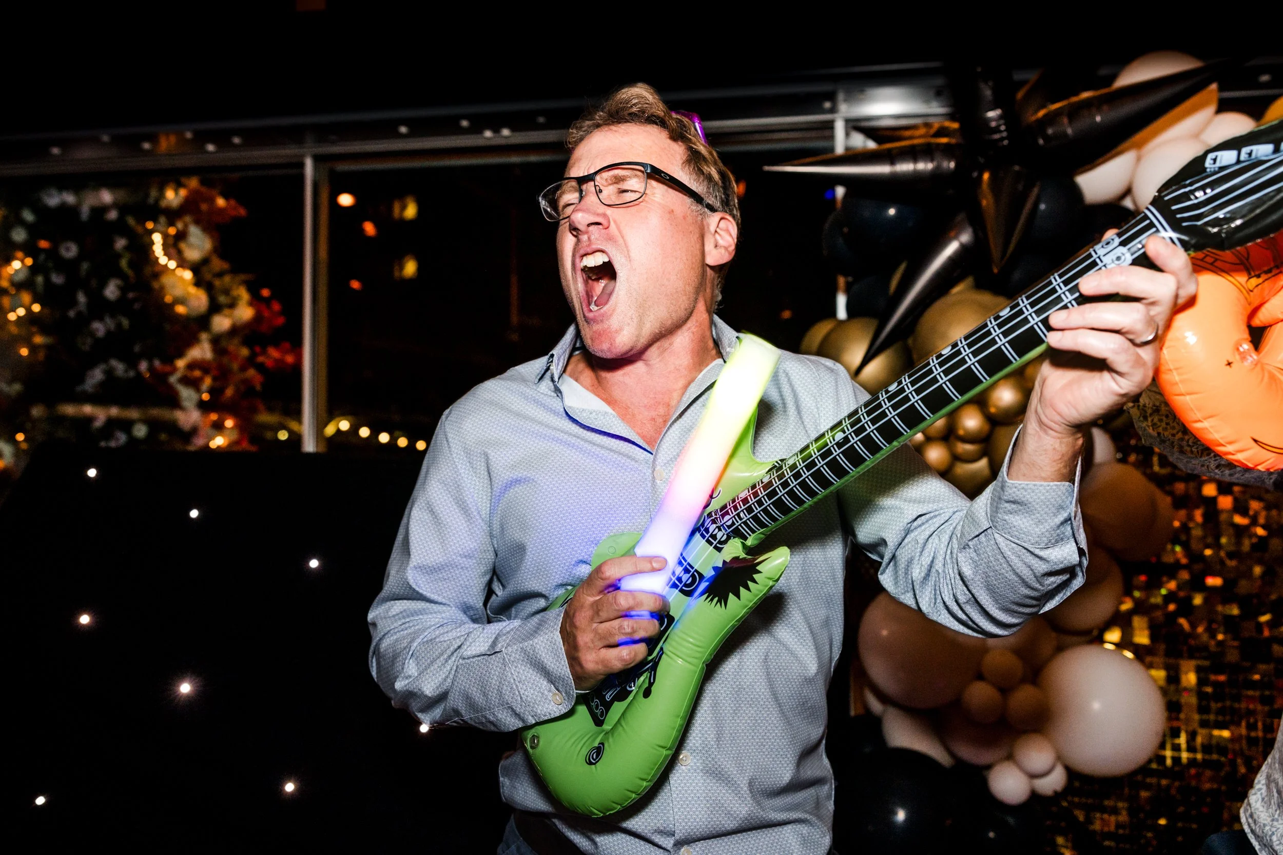 A man wearing glasses and a light-colored shirt is playing an inflatable green toy guitar at a party, singing passionately with his mouth wide open. Decorations with balloons and string lights are visible in the background.