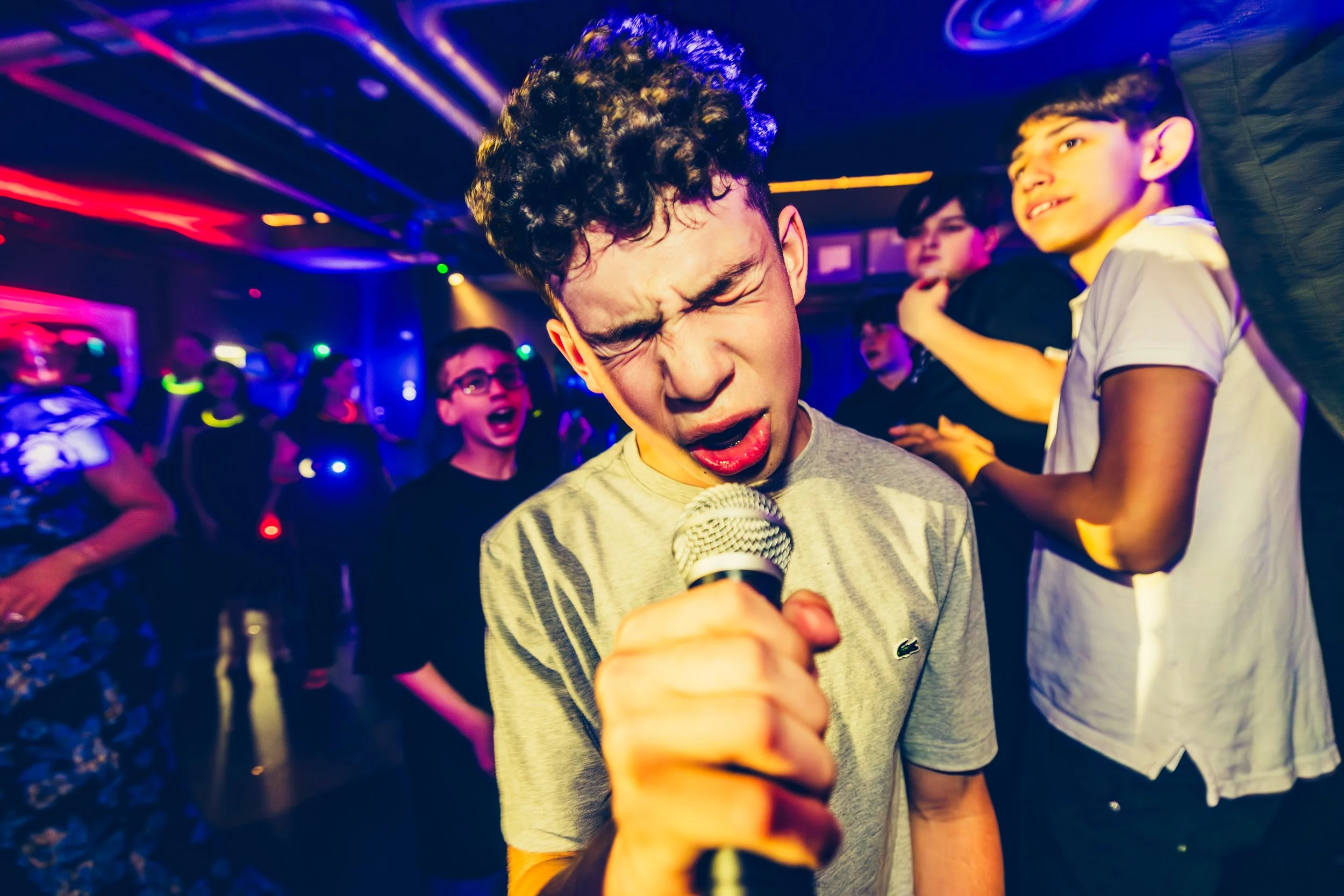 A young man passionately singing with a microphone at a luxury party, surrounded by other people dancing and enjoying the music, with colorful stage lights.