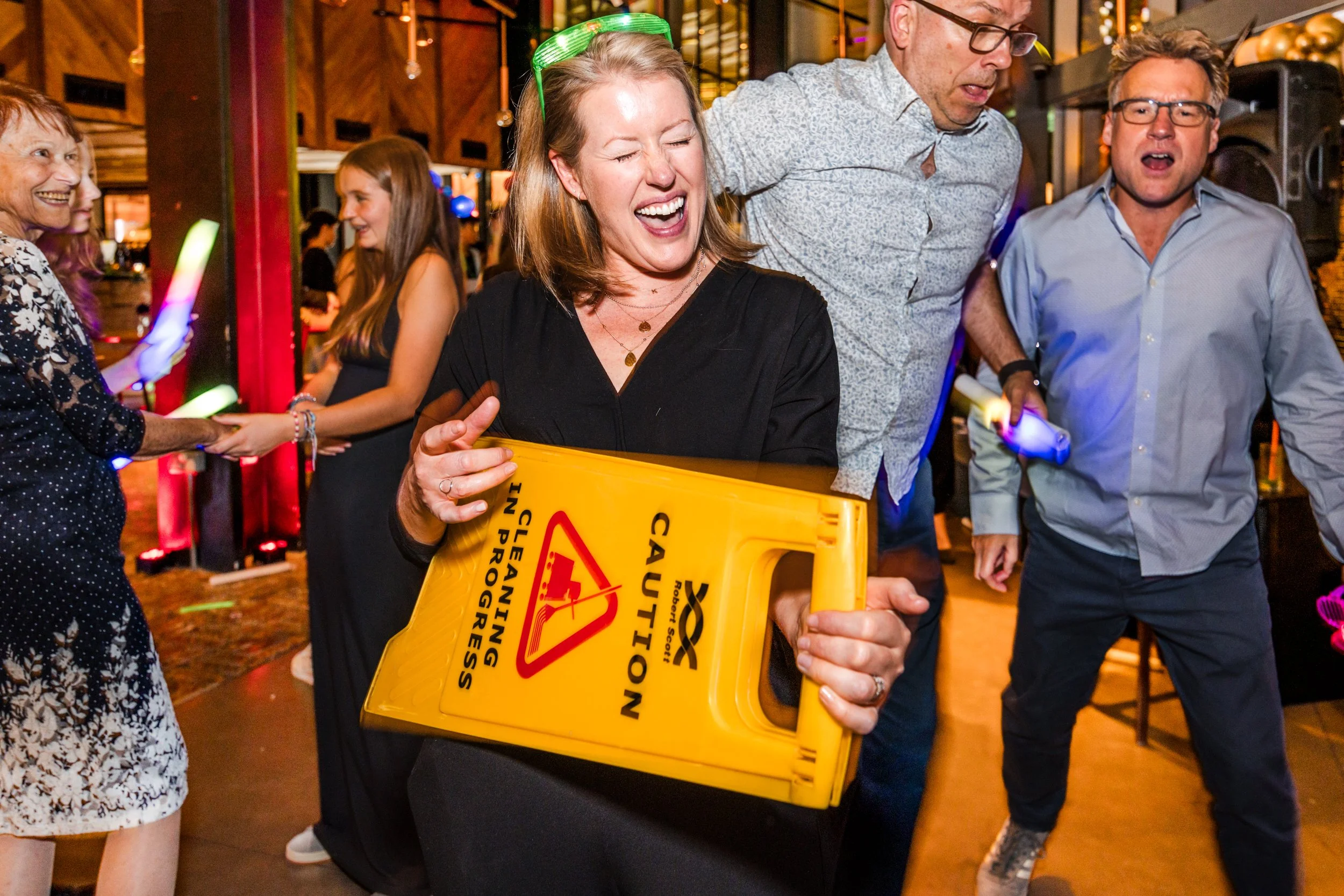 People at a simcha 
celebration, with a woman holding a yellow caution sign, while others dance and have fun in an indoor venue.