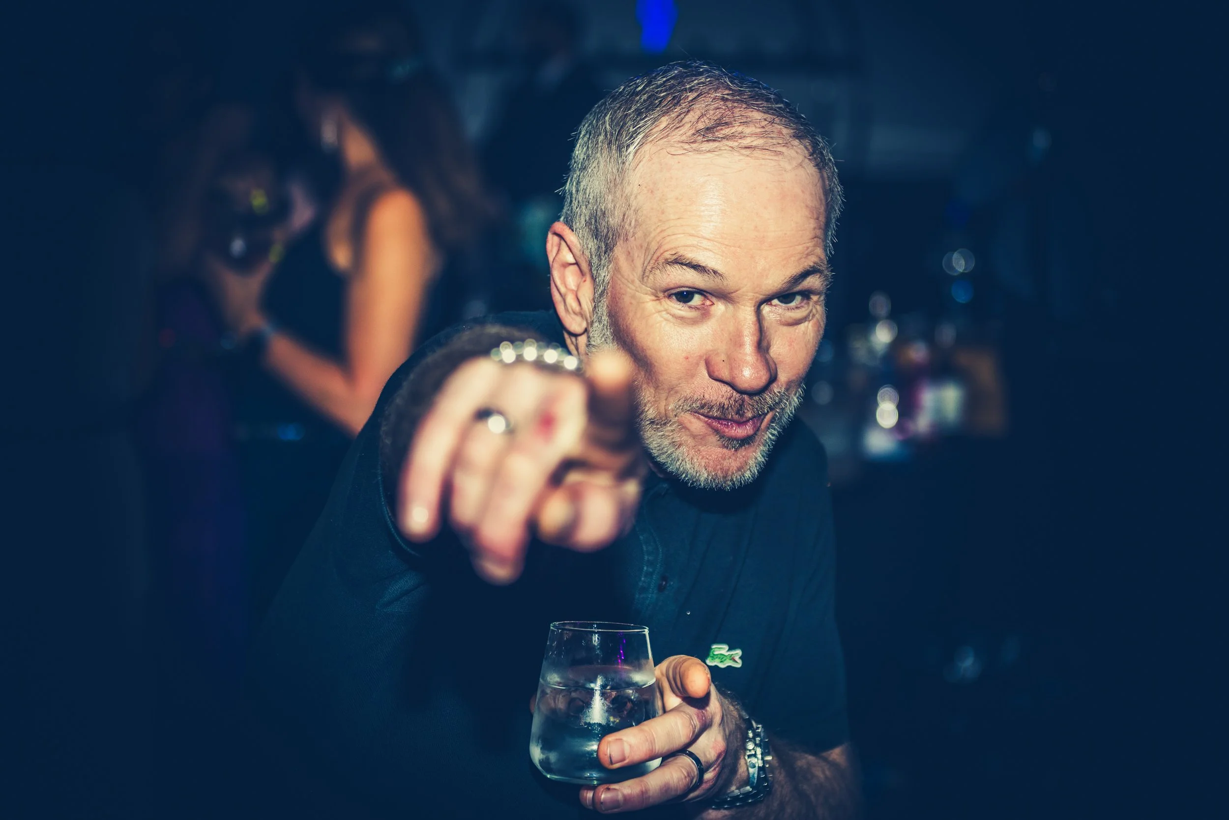 A middle-aged man with short gray hair and a beard pointing at the camera while holding a glass of clear drink at a luxurious party.