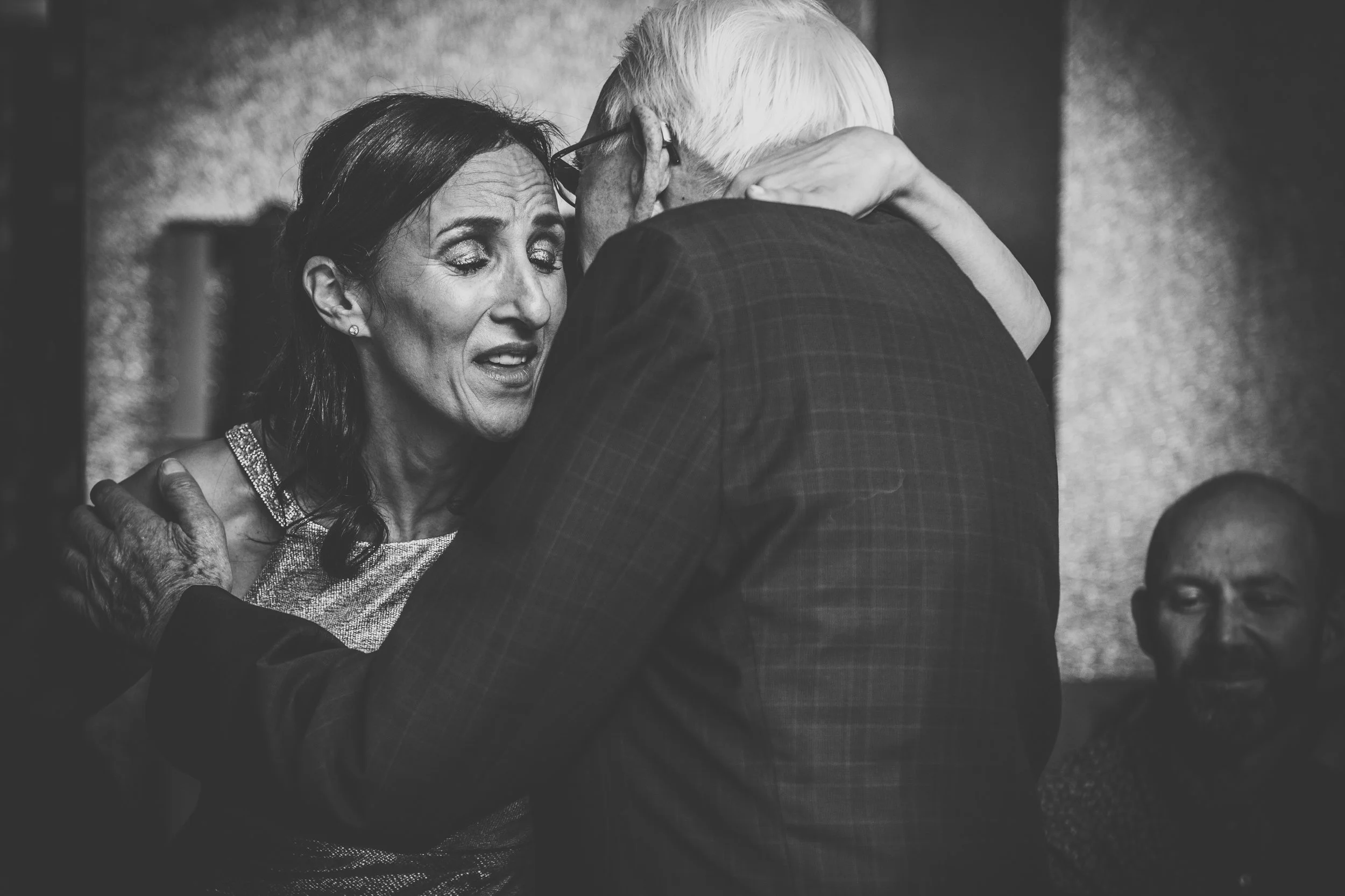 A grayscale photo of a woman embracing an elderly man, conveying a sense of emotion and connection. Another person is visible in the background.