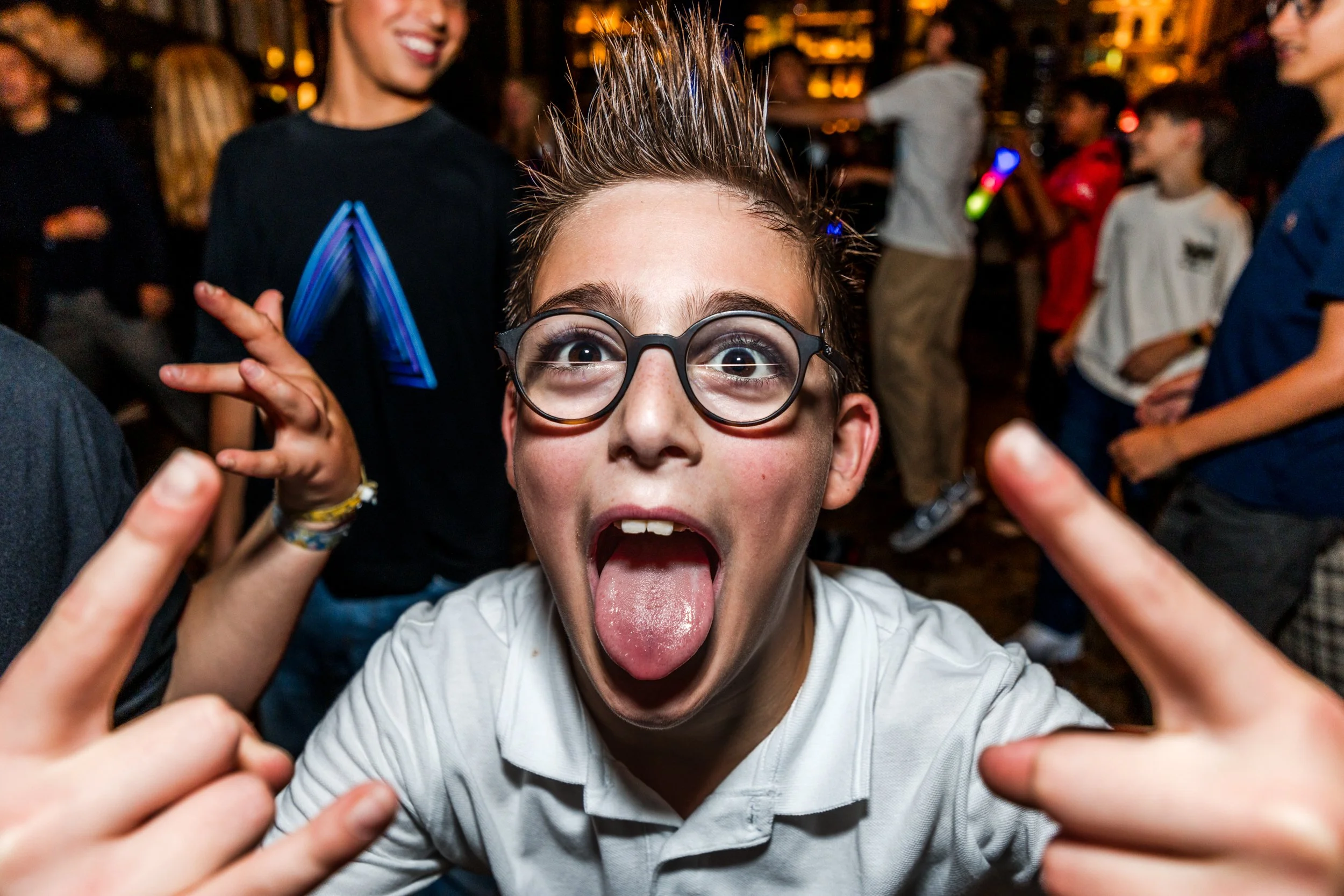 A young boy with glasses sticking out his tongue and making rock and roll hand gestures in a crowded social setting, at a bar mitzvah.