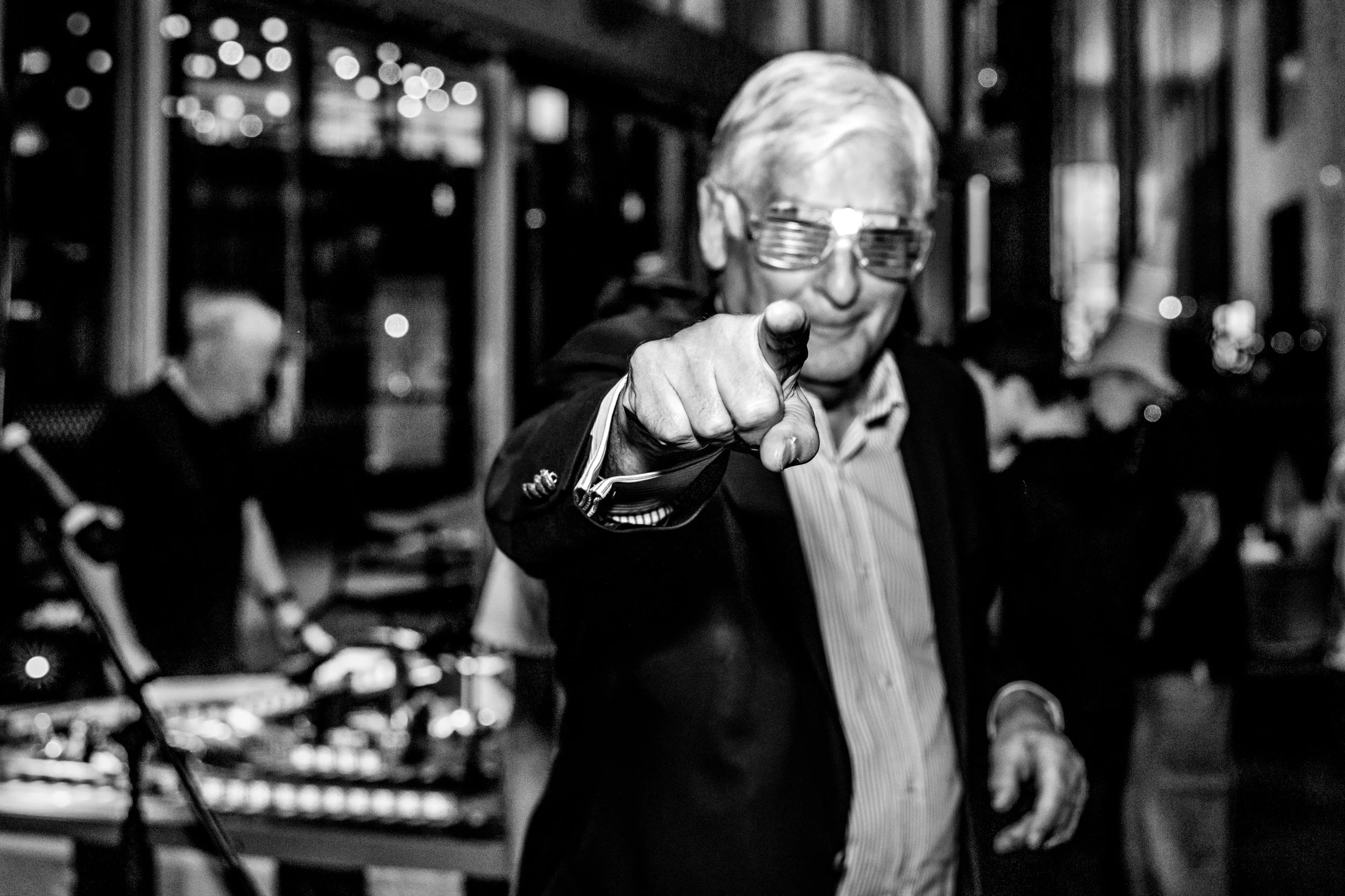 A man with gray hair and glasses pointing directly at the camera, smiling. At a bar mitzvah party, with other people and a musical instrument in the background.
