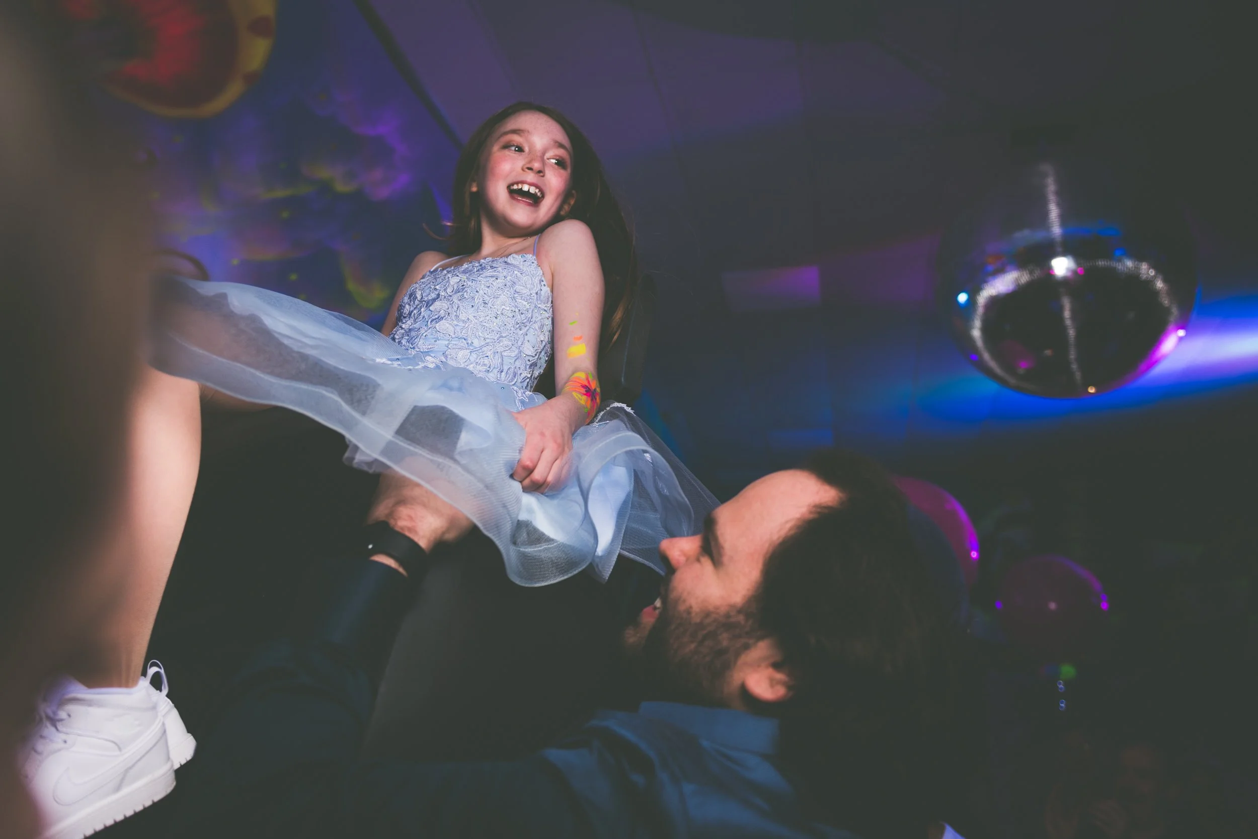 A young girl in a blue dress being lifted by a man at a bat mitzvah party with colourful lights and a disco ball in the background.