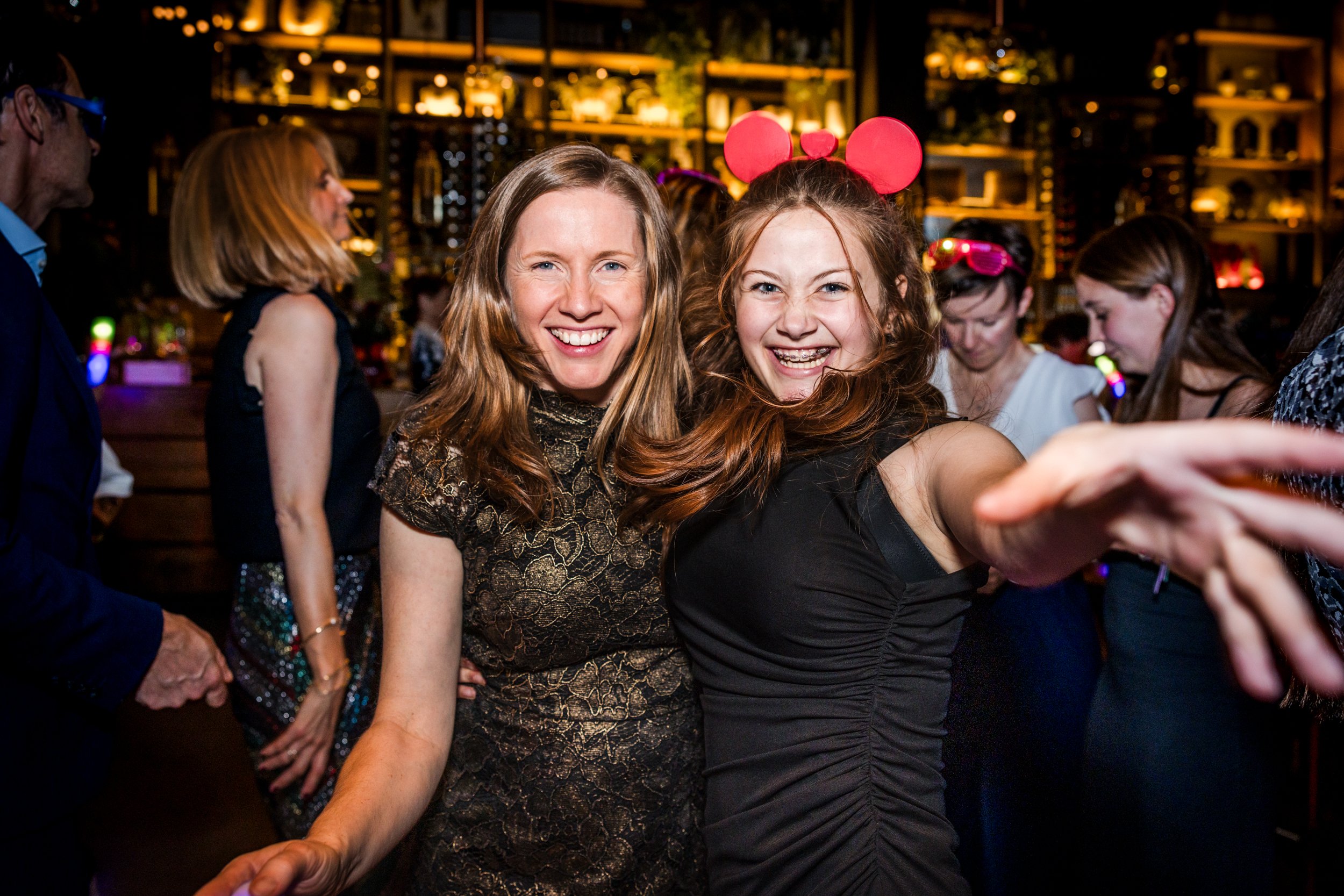 Two women smiling and taking a selfie at a luxury party with other people in the background.