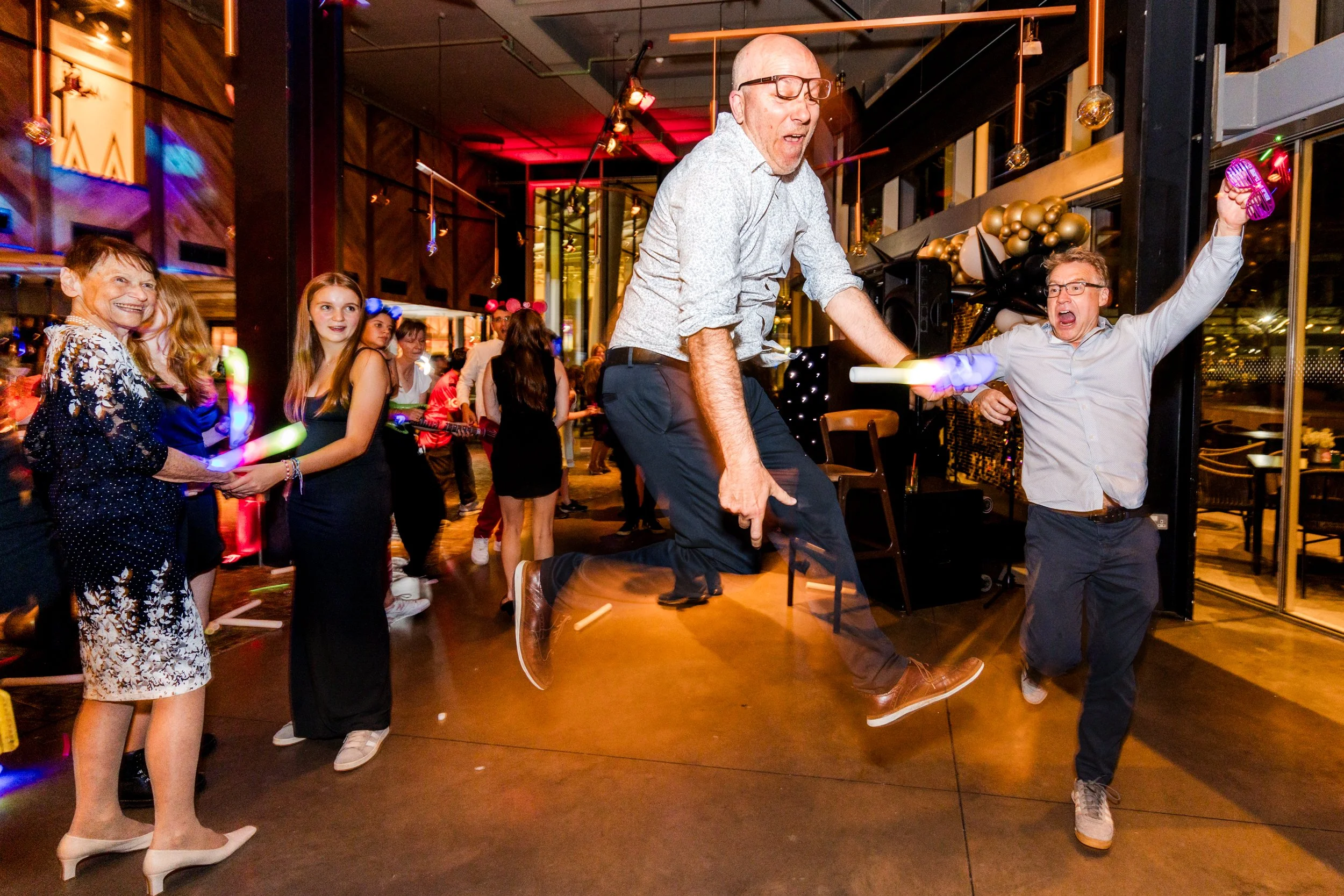 People dancing and celebrating at a party, with a man jumping in the air, surrounded by others smiling and holding glow sticks.