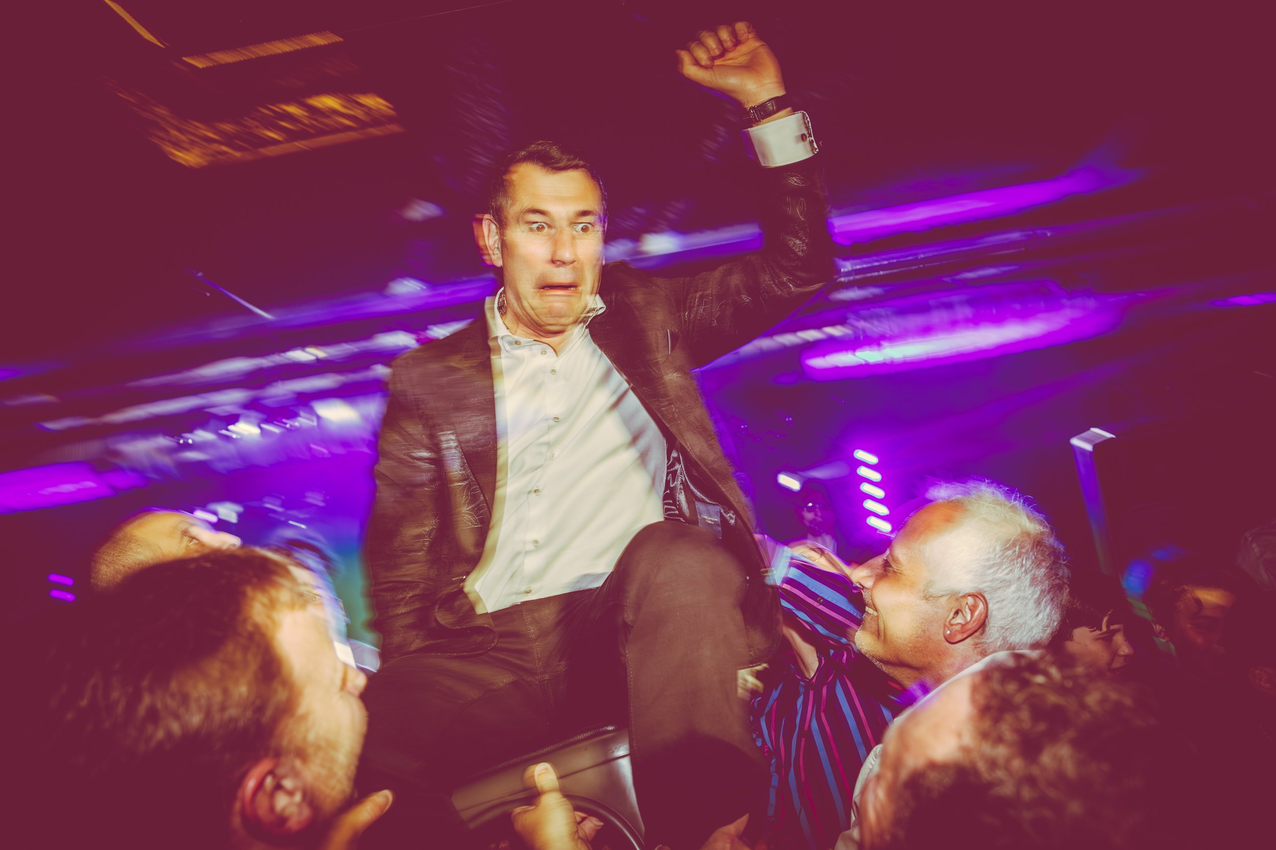 Man in a suit with a surprised expression being lifted by a group of smiling men at a bar mitzvah party with colorful lights.