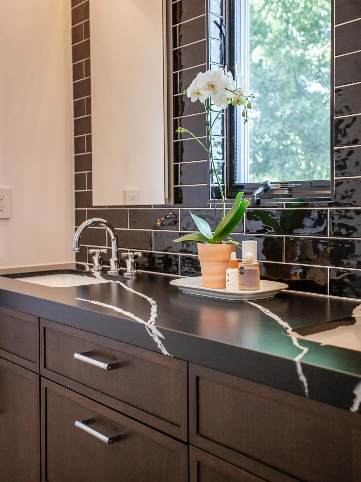 This primary bath design features a little bit of drama mixed in with classics. Its black and white palette is balanced with a rich wood vanity and cozy lighting. Gorgeous plumbing fixtures in classic polished chrome provide little details that spark