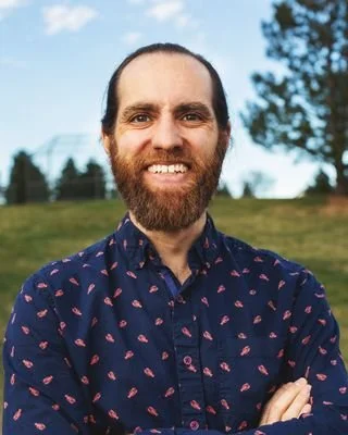 A woman with long dark hair, glasses, and a black top smiling at the camera against a plain background.