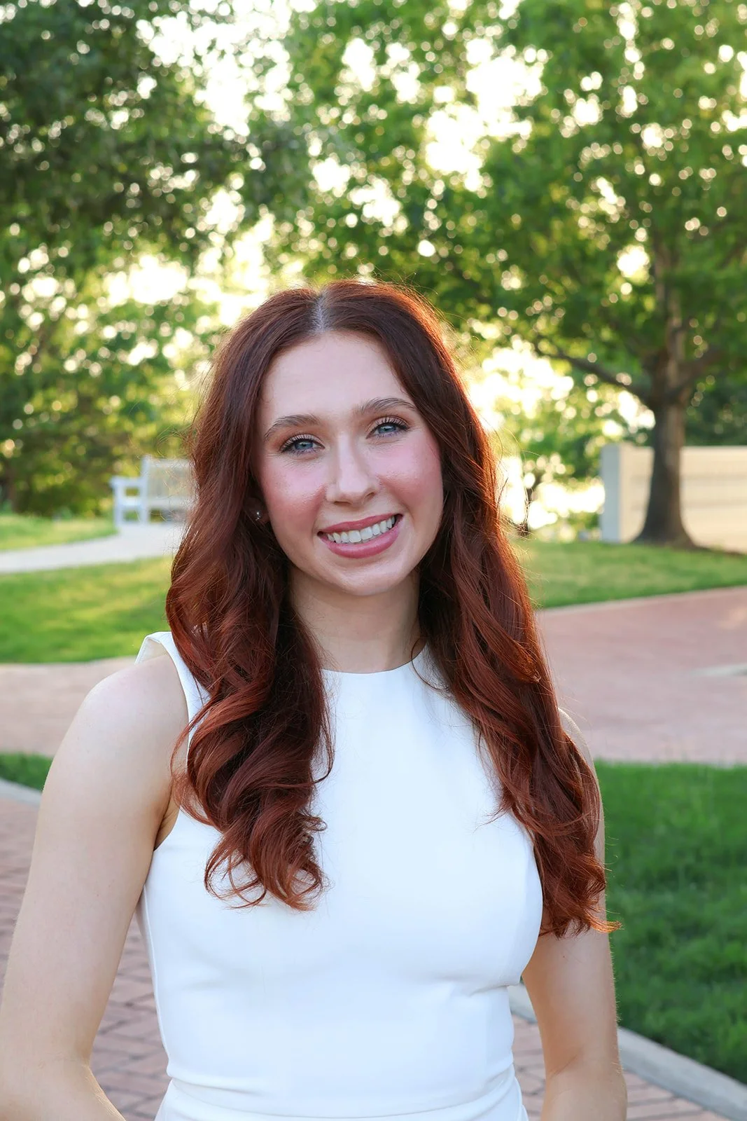 A young woman with red hair smiling outdoors in a white sleeveless top, with green trees and a paved path in the background.
