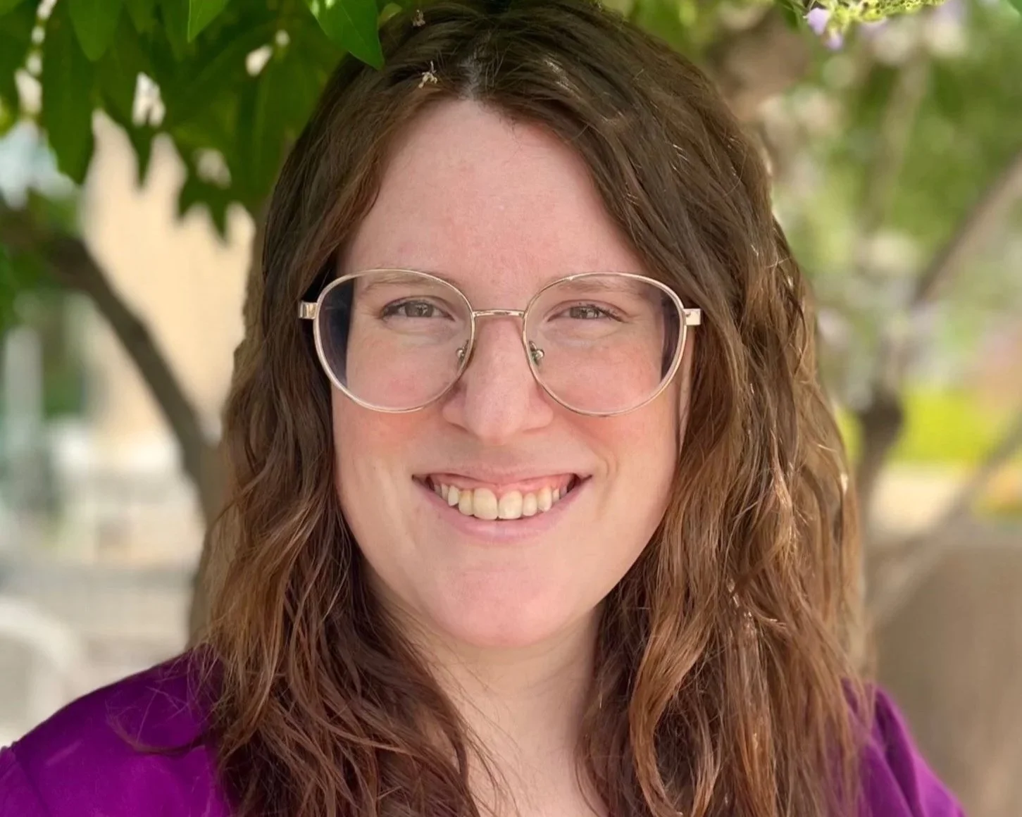 A woman with brown curly hair wearing clear glasses and a purple shirt, standing outdoors under green leaves, smiling at the camera.